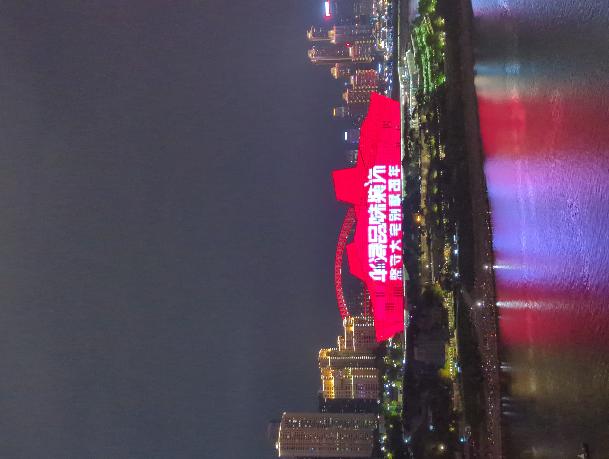 The image shows a nighttime cityscape in Yuzhong District, China. The most prominent feature is a large, red structure, possibly a building or bridge, illuminated with Chinese characters. The characters may represent a celebratory message or slogan. The red structure is situated along a riverfront, with reflections of the city lights and the red structure visible in the water. The riverbank is lined with greenery and pathways, suggestive of a park or promenade. Other buildings, including high-rises, are visible in the background, contributing to the urban skyline. Illumination from streetlights and buildings paints the scene.
