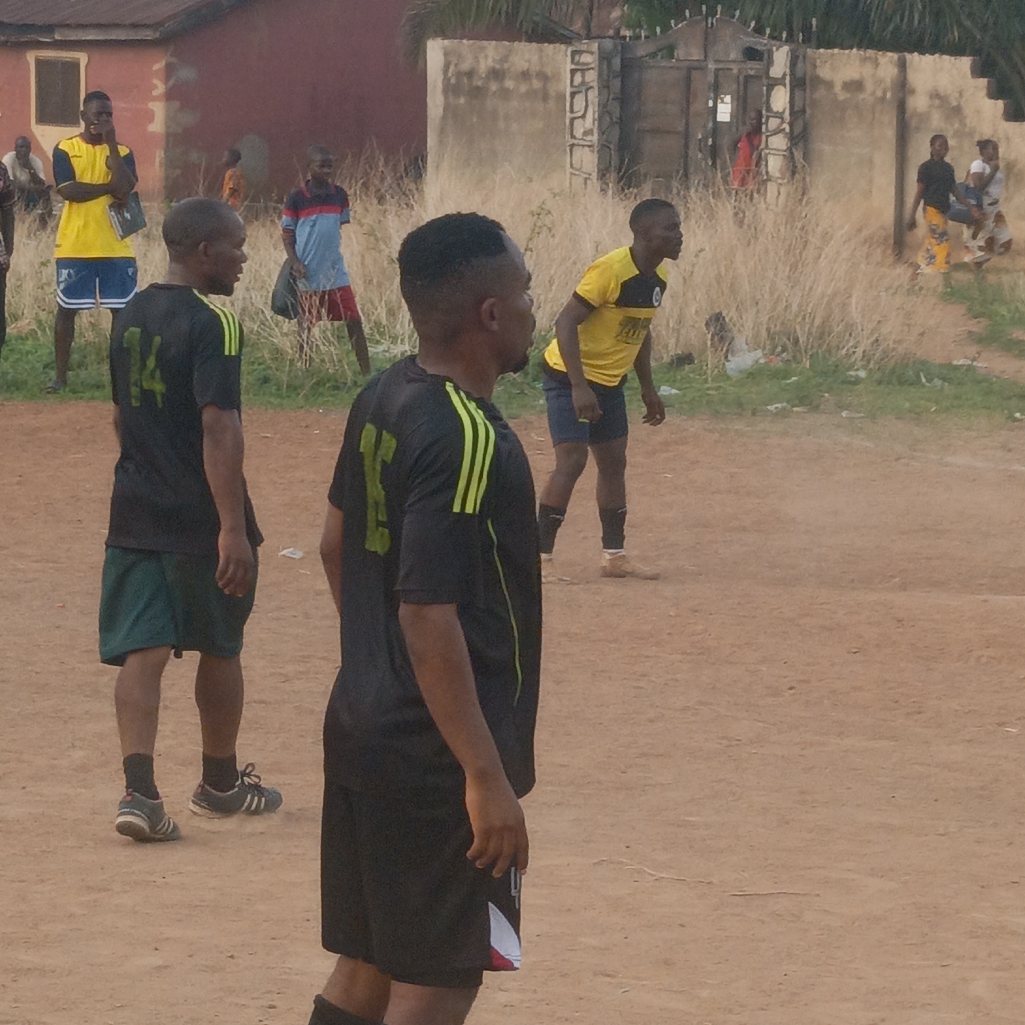 This outdoor scene, set in Akwanga, Nigeria, captures an informal football (soccer) game or practice session on a dirt field, likely in the late afternoon or early evening due to the warm, diffuse light.
The ground is predominantly reddish-brown dirt, with areas of tall, dry grass bordering the playing space. In the foreground, two men, facing away from the camera, wear black jerseys with bright green numbers and green stripes on the shoulders. The man on the left has "14" on his back and wears dark green shorts and black and white athletic shoes. The man on the right, slightly closer and in profile, has a partial "15" visible and wears black shorts.
In the midground, to the right, a man in a yellow and black jersey with dark blue shorts appears focused on the game, looking towards the right. To the left, another man in a yellow t-shirt and blue shorts stands with his arms crossed, holding a book, observing the activity.
The background features several onlookers and parts of buildings. A young man in a red and blue striped shirt stands near the dry grass. Further back, a person in a red shirt is visible inside a doorway of a building with a light-colored concrete wall. Two women can also be seen in the far right distance. The buildings range from rustic concrete structures to red-painted walls, typical of a semi-urban or rural Nigerian setting. The overall mood is relaxed and community-oriented, centered around the casual sporting event.