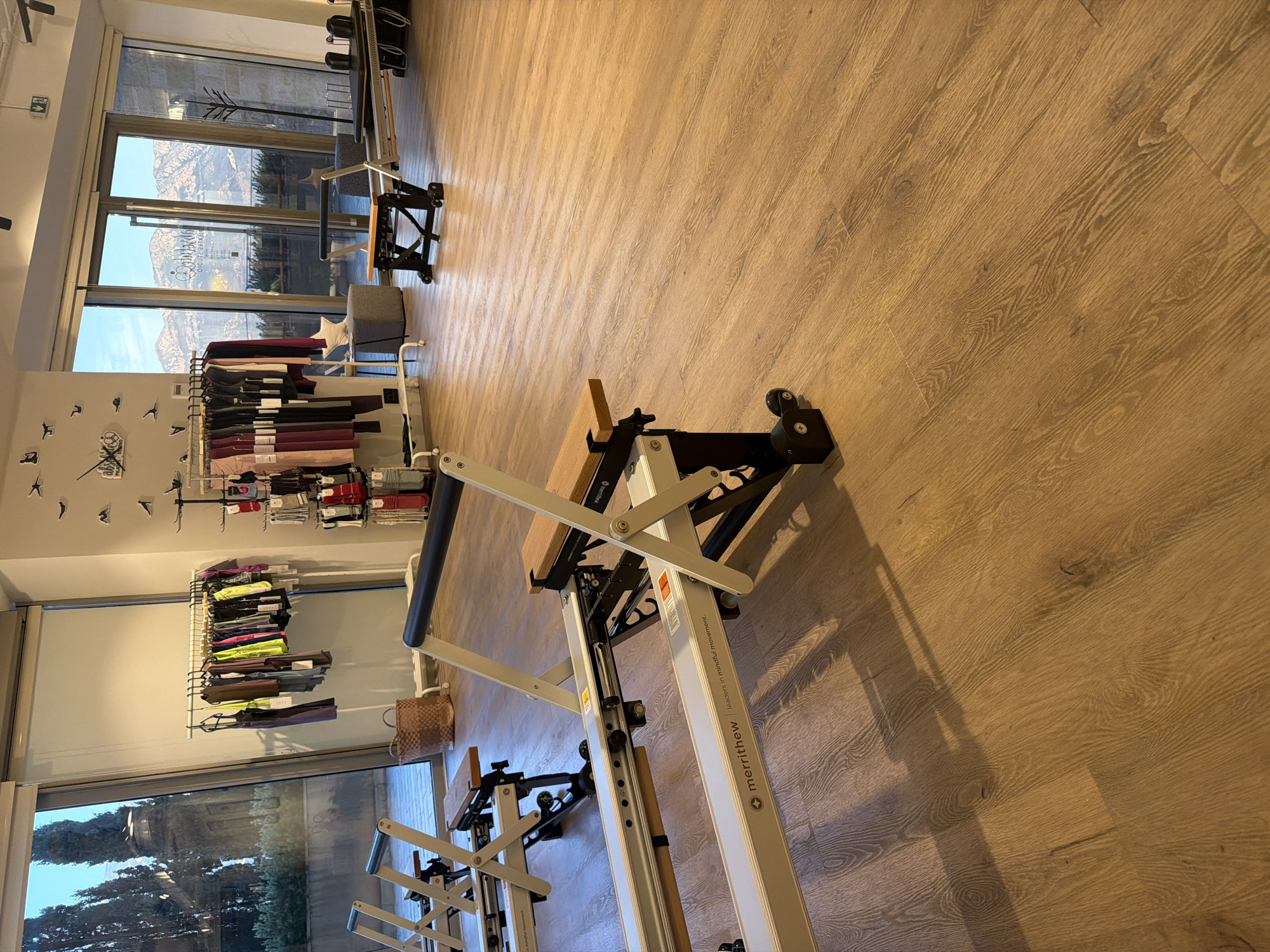 The image depicts the interior of a Pilates studio in Boreti, Montenegro. The space features a wooden floor and large windows offering views of trees and mountains. Several Pilates reformers, a type of exercise equipment, are arranged throughout the room. In the background, there is a rack of athletic clothing for sale. The studio appears clean and well-lit, suggesting it is operational and ready for use.