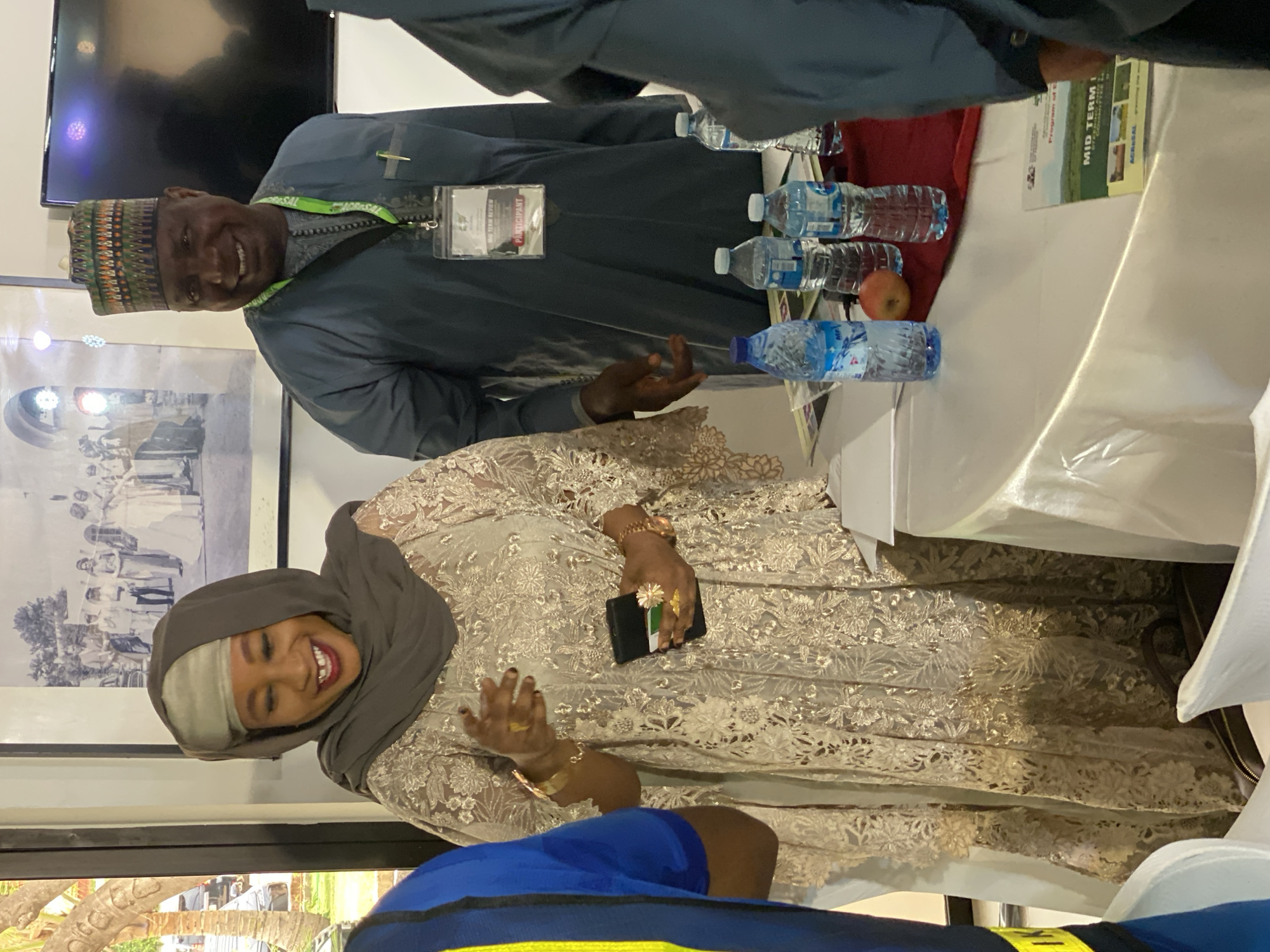 Two individuals are present in an indoor setting in what appears to be Kwarau, Nigeria. One is a woman wearing a hijab and an ornate, lace-like garment. She is smiling. The other person is a man wearing a traditional hat and a garment, with a name tag visible on his chest. They are positioned around a table covered with a white tablecloth, on which several bottles of water and a red apple are placed. A booklet or program with the words "MID TERM" on its cover is also on the table. In the background is a black-and-white framed photo.