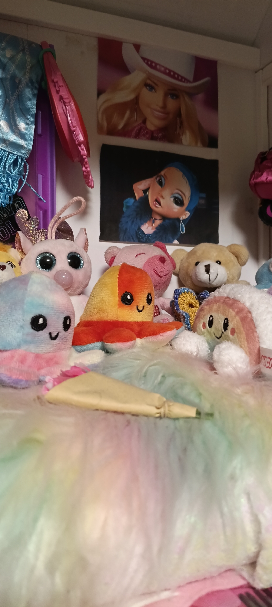 This is a close-up, eye-level, indoor shot of a collection of stuffed toys. The toys are in various colors and styles, including an octopus-like creature with a blue and pink tie-dye pattern, another octopus-like creature with orange and yellow segments, a fluffy white teddy bear, and a pink pig with large blue sequined eyes. In the background, there are two posters of animated characters. One poster depicts a blonde woman wearing a white cowboy hat, and the other shows a young woman with blue hair and blue eye makeup. There are also glimpses of doll accessories in the upper left corner, including a blue beaded scarf and a pink braided accessory. The overall scene is colorful and playful, suggesting a child's bedroom or play area. The location of Wels, Austria, is not visually represented in this image.