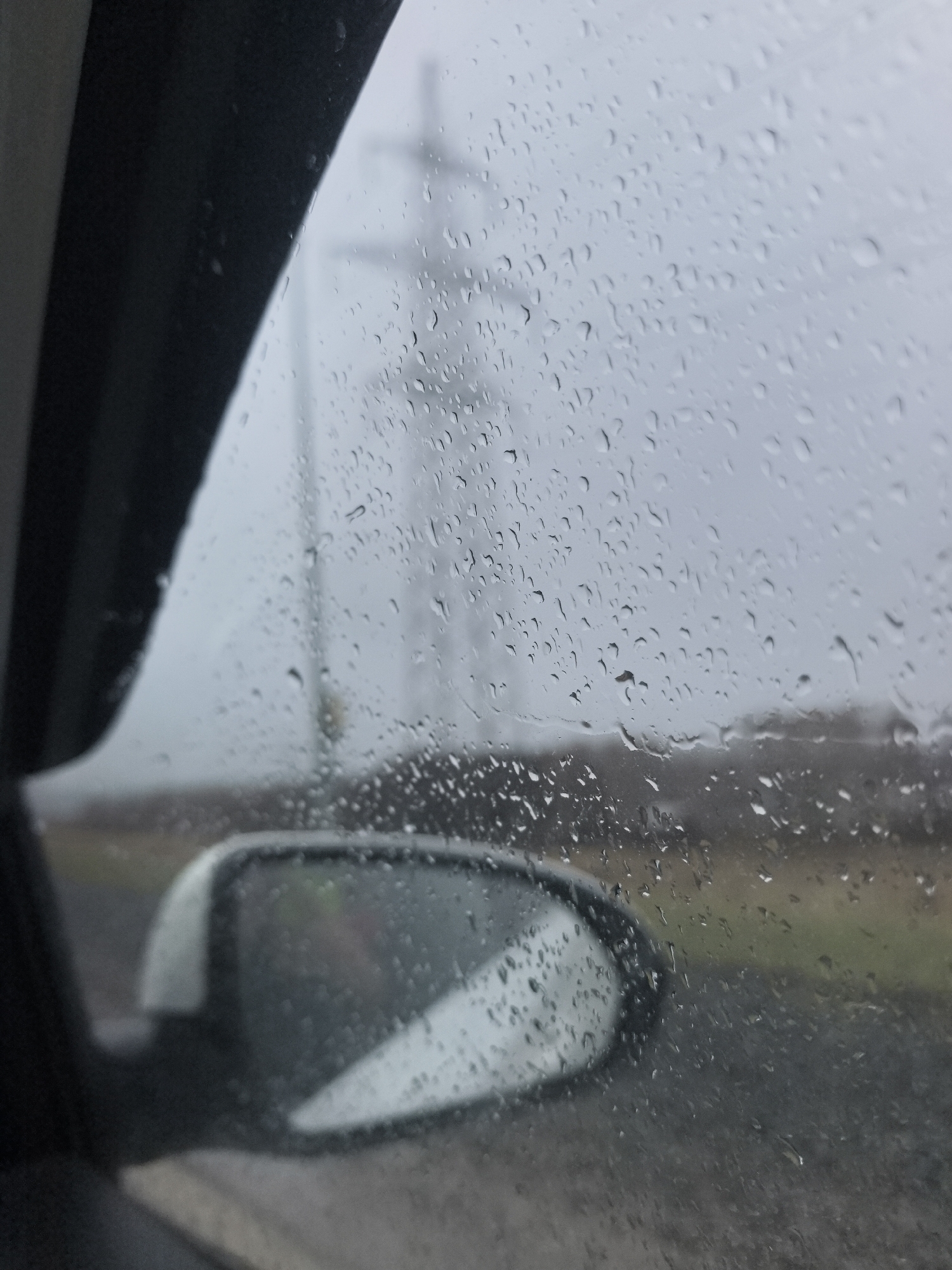 The image presents a somber view from inside a vehicle, looking out a rain-streaked side window on an overcast day. The left side of the frame is occupied by the dark, curved interior panel of the car, with the window filling the majority of the composition. Numerous water droplets of various sizes cling to the glass, creating a textured, diffused effect.
Through the rain-covered window, an external scene unfolds with blurred details. In the lower right, the car's side mirror is visible, also dotted with raindrops, reflecting a distorted and indistinct view of the immediate surroundings. The landscape beyond the vehicle appears hazy and muted in color, indicative of low visibility due to rain or mist.
In the background, a large, dark, cross-shaped structure, likely a power line pylon or electricity pole, stands against the grey sky. Below, a strip of muted green and brown landscape suggests distant fields or undeveloped land bordering what appears to be a road or path. The overall lighting is dim, reflecting the gloomy weather conditions of a rainy, cloudy day. No specific activity is depicted other than observing the weather from inside the car. There is no visible text in the image. The setting suggests a roadside or rural area, consistent with a location like Noviy, Russia, during a wet, overcast period.
