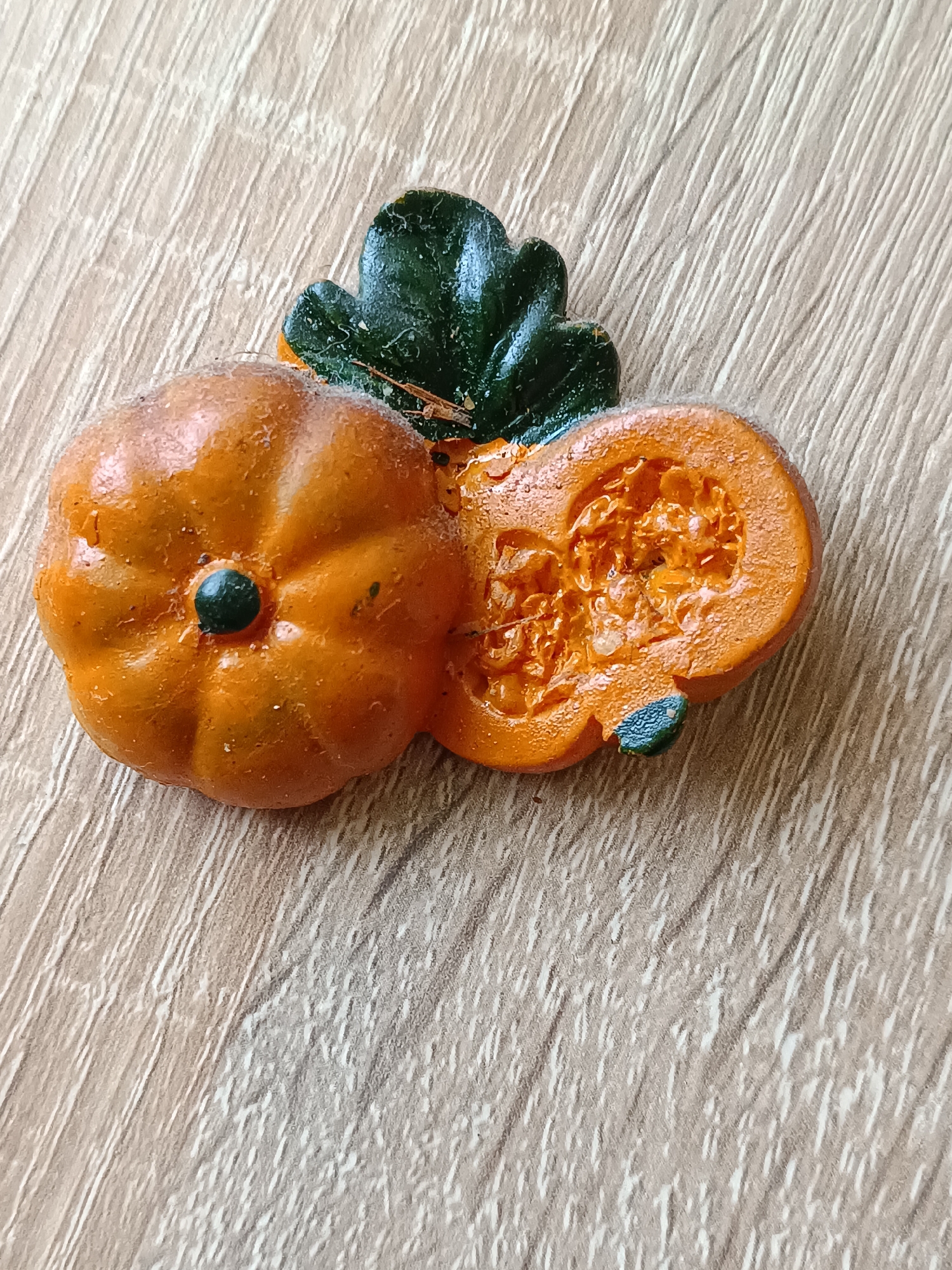 A close-up, overhead shot shows a faux pumpkin decoration with a leaf. The pumpkin is orange and appears to be cut in half, revealing a textured orange interior with seeds. A dark green leaf is positioned behind the pumpkin. The decoration rests on a light brown wooden surface with a visible wood grain pattern. The location is not specified within the image.