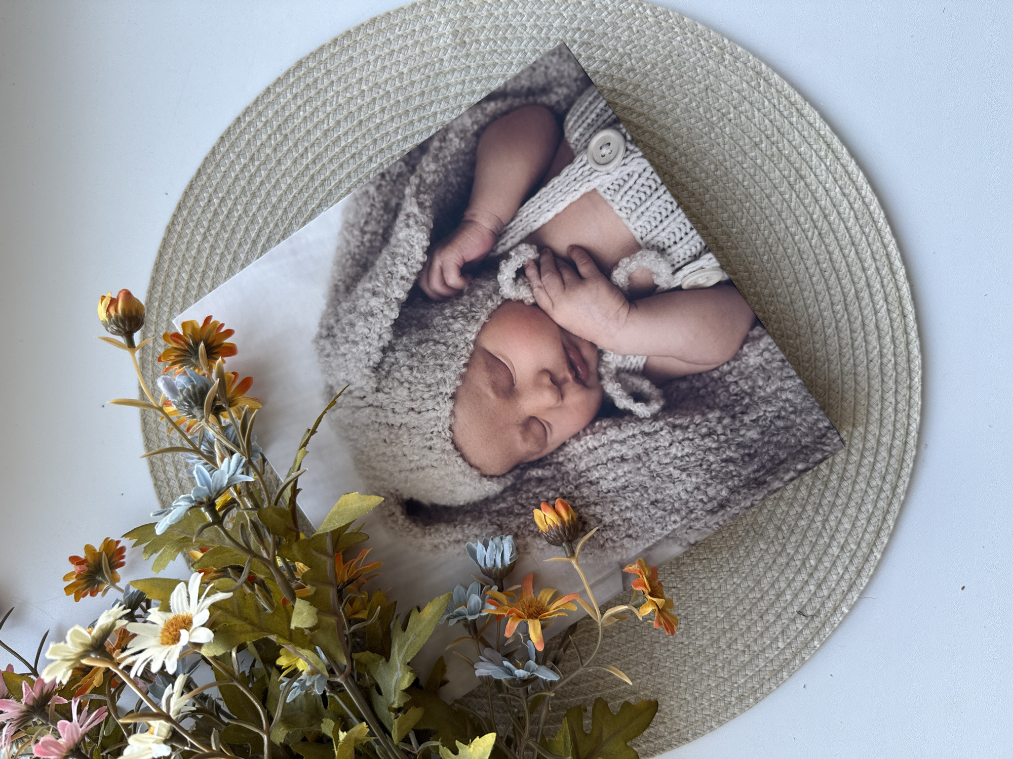 A photograph depicts a newborn baby sleeping, swaddled in a gray knitted outfit, and a matching bonnet. The baby is lying on a white surface, with a round beige woven placemat underneath the picture. A bouquet of artificial daisies with multi-colored petals is positioned on the left side of the photo. The scene appears to be set indoors on a white table. There is no information to suggest a location in the city of Rusanovo, Russia.