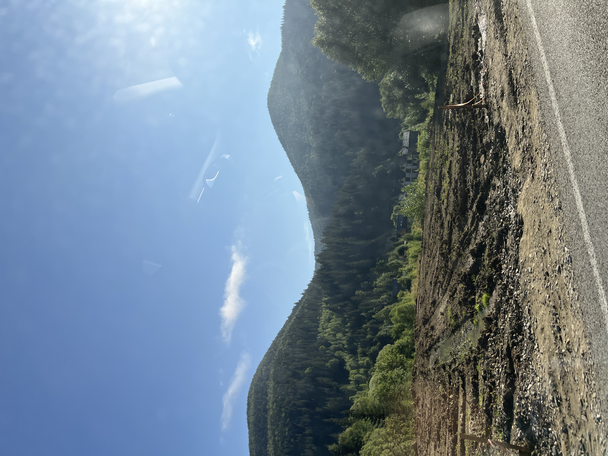 This image depicts a rural landscape with mountains covered in dense green forests under a clear blue sky. In the foreground, a muddy, disturbed field shows signs of recent work, with tire tracks and small puddles of water. There are some scattered rocks and a few wooden stakes visible in the field. In the middle ground, nestled among trees, are several buildings with dark roofs. The scene appears to be a view from a roadside, as a portion of an asphalt road with a white line is visible in the bottom right corner. The location is identified as Tatariv, Ukraine.