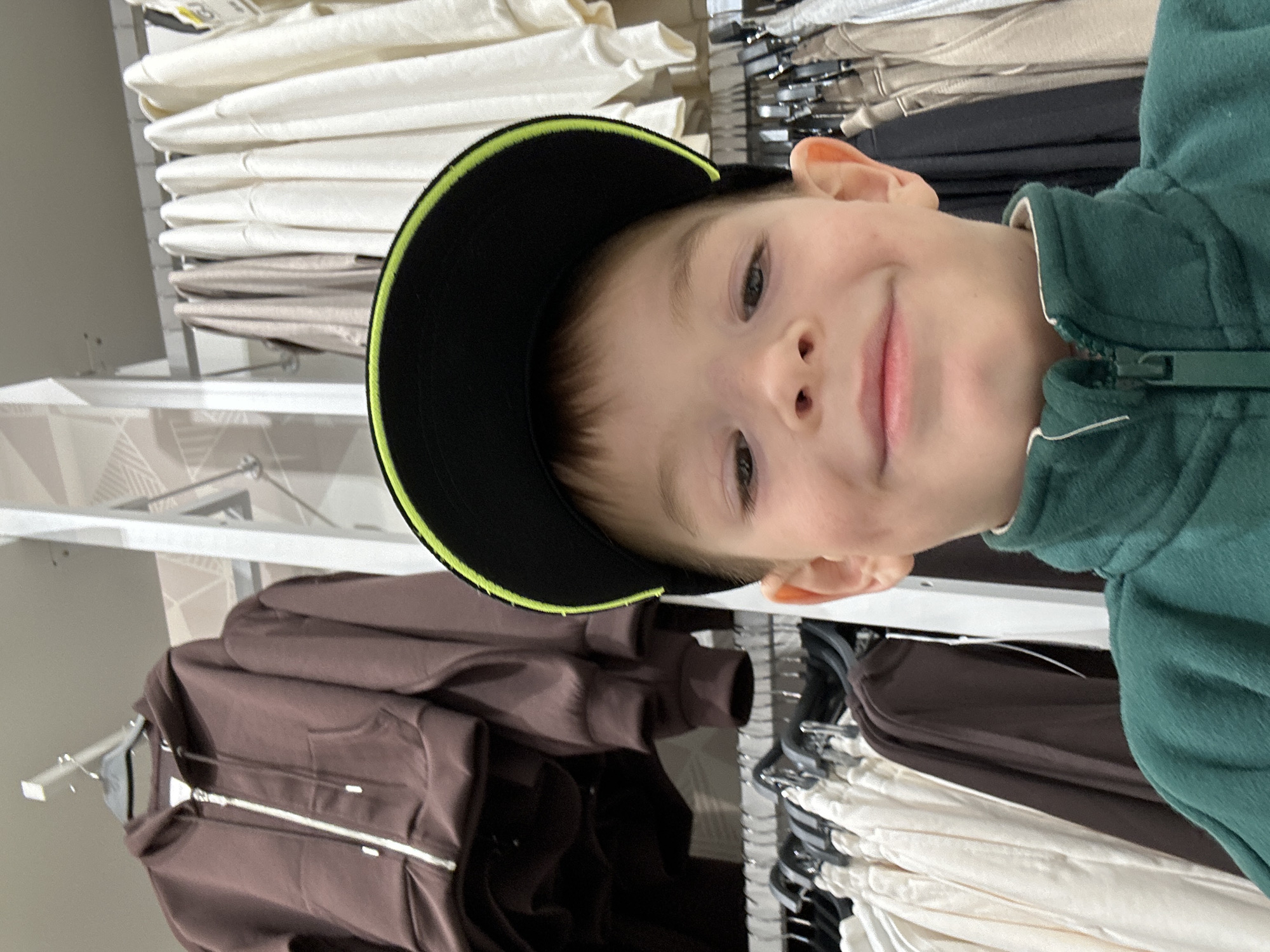 A young boy wearing a green zip-up jacket and a black baseball cap with a green brim is posing for a selfie in a store. The background shows racks of folded clothing, including white, beige, and brown items. The location appears to be a clothing store in a city that may be Chaikovsky, Russia.
