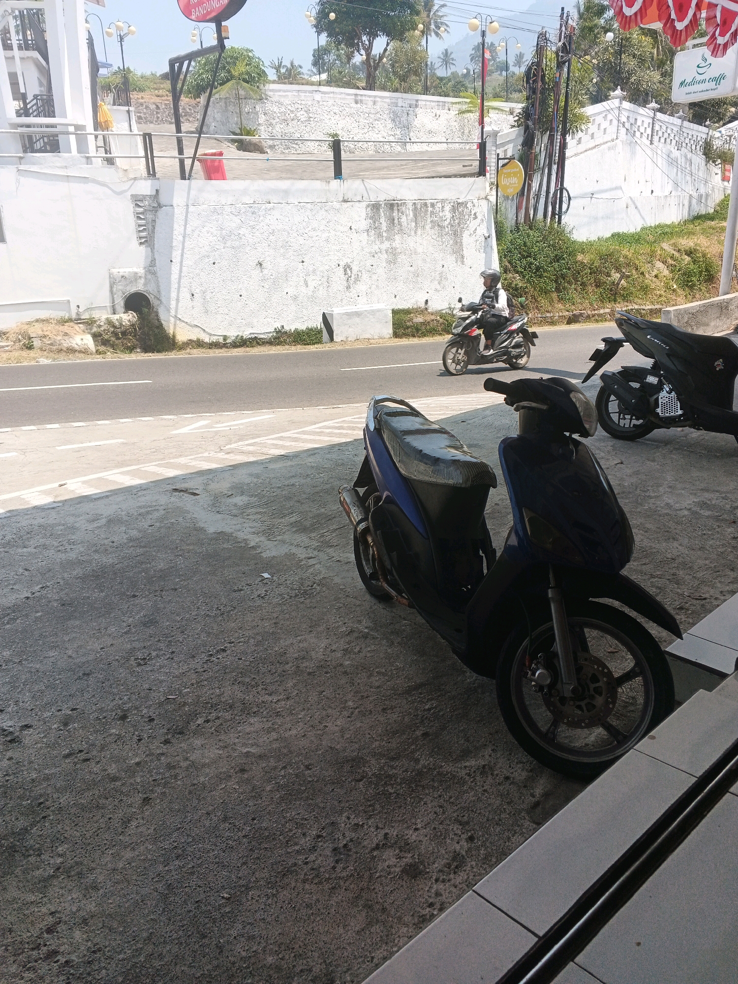 In Pakopen, Indonesia, a blue scooter is parked on a concrete surface in front of a building. The scooter is facing left, with its front wheel clearly visible. Behind the blue scooter, a black scooter is also parked, facing right. In the background, a red and white banner hangs from a building. A person on a red and black scooter rides on the asphalt road. The road has white markings, indicating lanes. The environment appears to be outdoors, with trees and a white wall in the distance. The sky is visible, suggesting it is daytime.