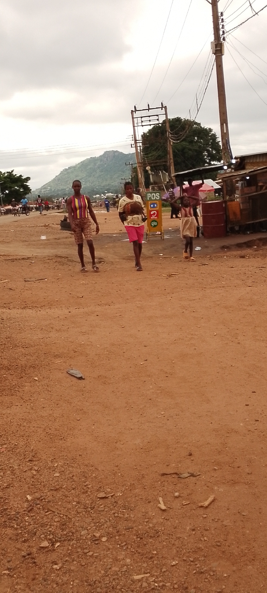 In Gboko, Nigeria, on a dusty road, three individuals are walking. In the foreground, a person in a striped shirt and patterned shorts walks from left to right, followed by another person in a light-colored shirt and pink shorts carrying a brown object. Further back, a young girl in a light-colored dress walks. In the background, there are people on motorcycles, a POS sign, and a building with corrugated metal roofing. A mountain is visible in the distance under a cloudy sky. The ground is covered in dirt and small stones.