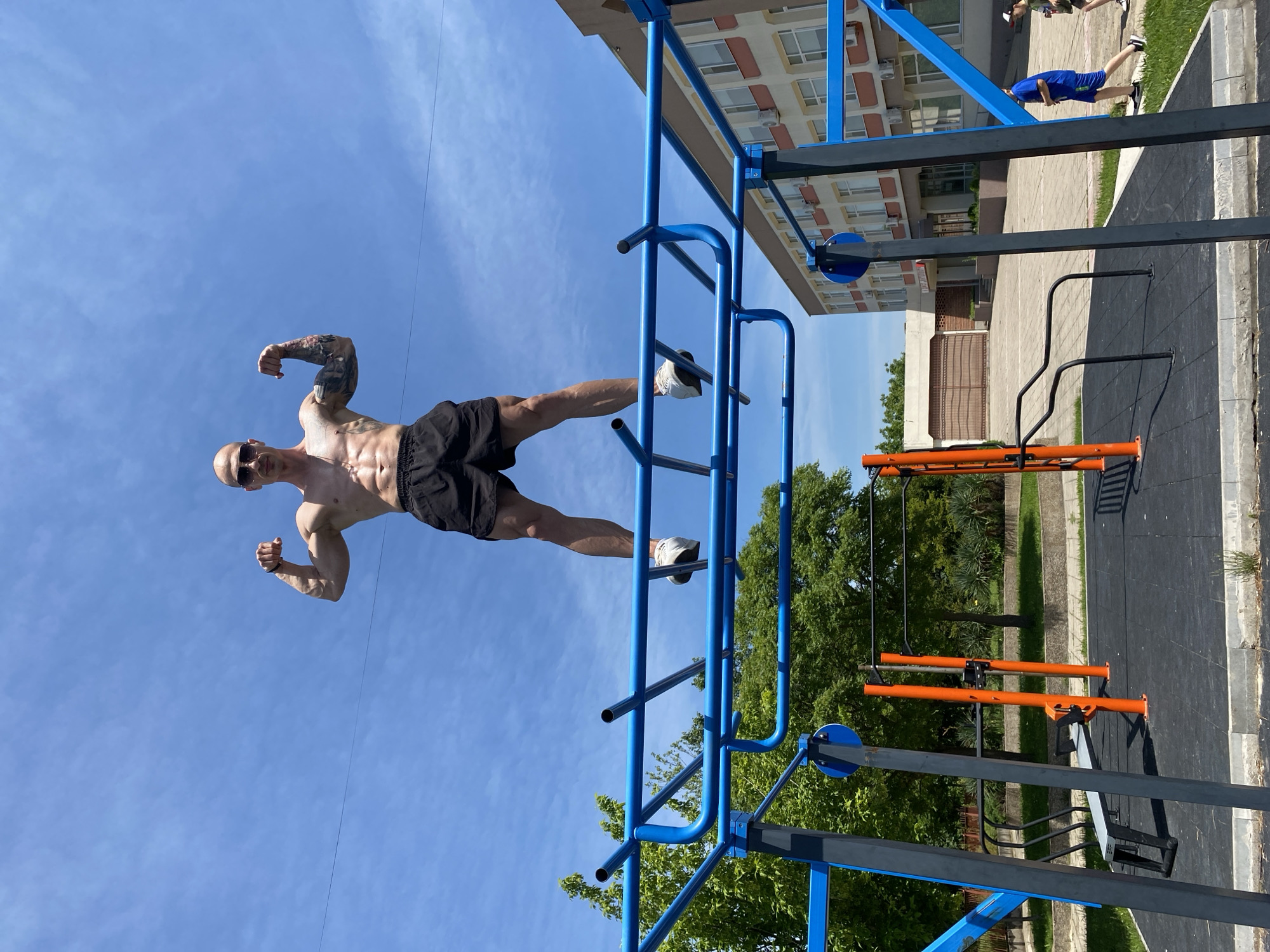 A fit, bald man wearing sunglasses and black shorts is performing a full planche on a blue and gray outdoor exercise structure, likely in a park. He is facing toward the left with his arms flexed and legs extended horizontally. Tattoos are visible on his arms. His feet are clad in white and gray sneakers. Behind the exercise structure, a building with multiple windows and balconies is visible, along with trees and other exercise equipment in orange and gray. Another person in a blue shirt can be seen in the upper right corner of the image. The sky is blue with very few clouds. The location is Pleven, Bulgaria.