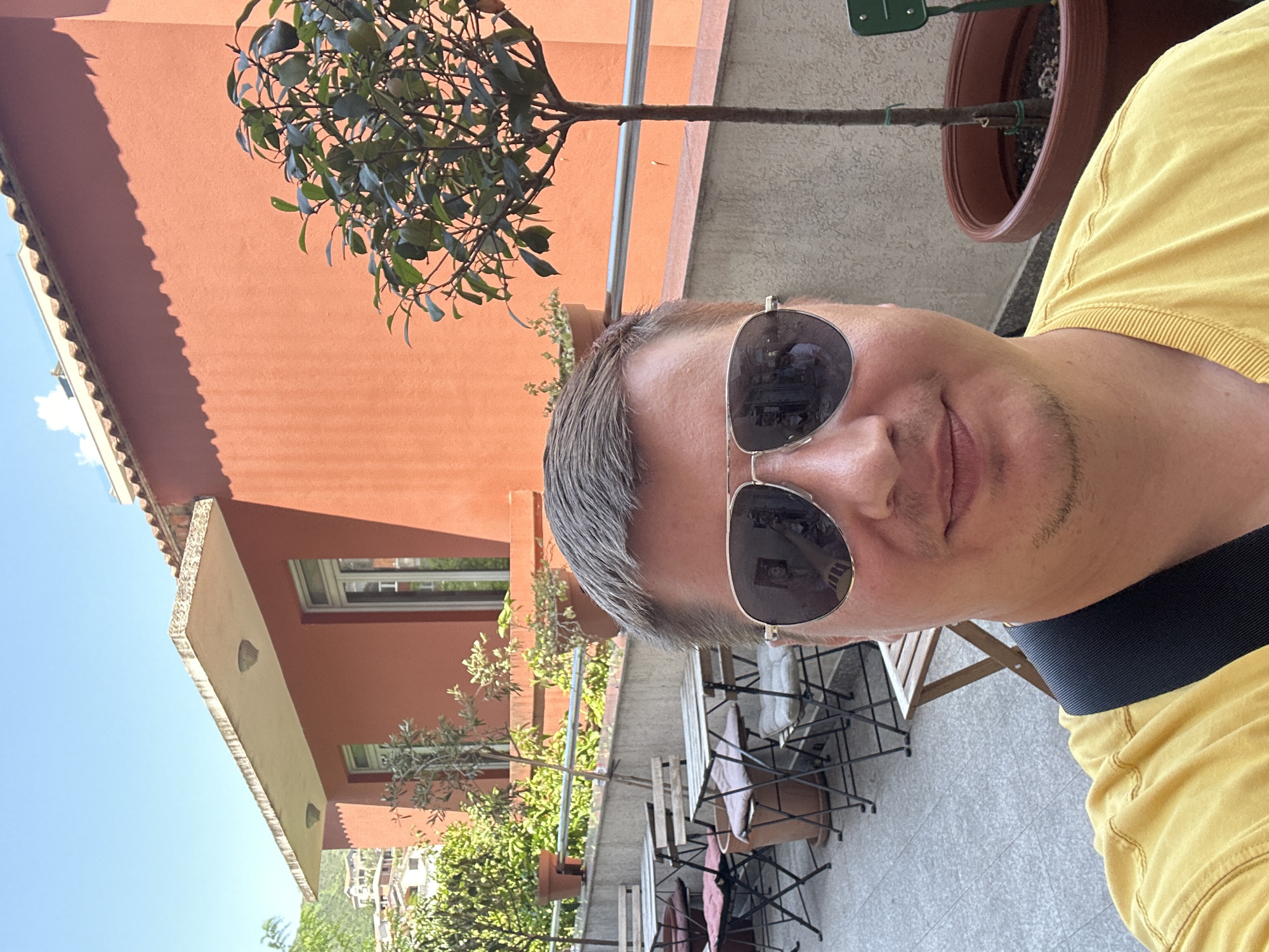 This is a selfie taken outdoors on a sunny day, likely in the afternoon, given the bright light and shadows. The subject is a person with short, graying hair and sunglasses, wearing a yellow shirt. They are standing on a patio or balcony with a tiled floor. In the background, there's an orange building, a small tree in a pot, and more greenery. Further in the distance, a hilly landscape with buildings is visible, suggesting a Mediterranean or coastal location. There are also a few outdoor tables and chairs visible, indicating a seating area. The overall mood is relaxed and casual, consistent with a vacation or leisure setting. The clear blue sky and sunlight suggest good weather.
