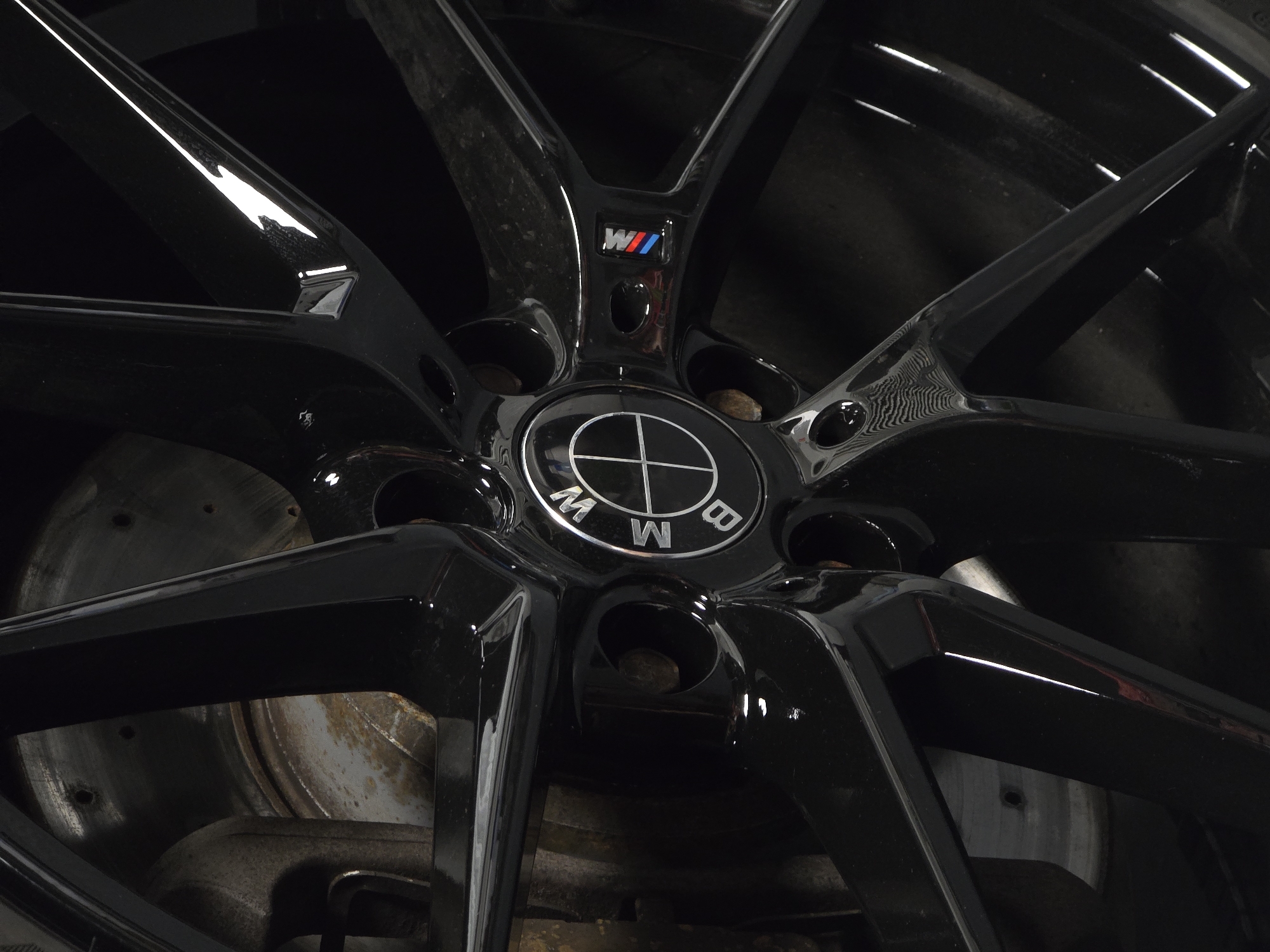 A close-up, low-angle shot captures a black BMW M-Sport wheel and brake rotor. The wheel features a multi-spoke design with a glossy black finish that reflects light. The center cap displays the BMW logo. To the right of the center cap, a smaller M-Sport badge with red, white, and blue stripes is visible. The brake rotor behind the wheel appears somewhat weathered and rusty. The background is dark and out of focus, suggesting an indoor or dimly lit environment. The image does not depict any people or specific landmarks of Ruse, Bulgaria.