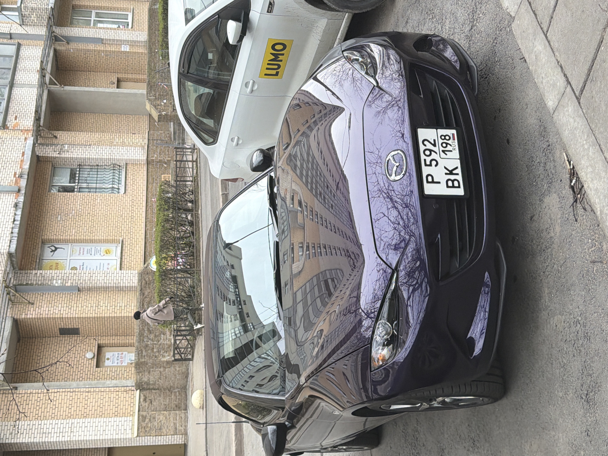 The image captures a street scene in Saint Petersburg, Russia, featuring two cars parked alongside a multi-story brick building.
In the foreground, a dark purple Mazda sports car is prominently displayed, positioned at an angle on the asphalt road. Its glossy surface reflects the surrounding urban environment, including the brick buildings and bare tree branches, suggesting a season like late autumn, winter, or early spring. The car has a Russian license plate, which reads "P 592 BK 198 RUS", with "198" specifically identifying the Saint Petersburg region.
Partially visible behind the Mazda is a white Volkswagen Polo sedan. A large yellow rectangular sticker with the word "LUMO" in black letters is affixed to its side door, indicating it might belong to a car-sharing or delivery service.
The background consists of a light brown brick building, characteristic of urban architecture. It features multiple windows, some with security grilles, and balconies. In the middle ground, near the building's entrance, a person is visible walking or standing on the sidewalk, dressed in a light-colored coat.
Visible text includes the car registration details on the Mazda's license plate and the "LUMO" branding on the white car. Signs are also present on the building; one on an upper window appears to show a stylized figure and some Cyrillic text, possibly related to a business like a studio, while another sign on a lower door contains blurry text and numbers. The scene appears to be taking place during daylight hours, possibly under overcast conditions.