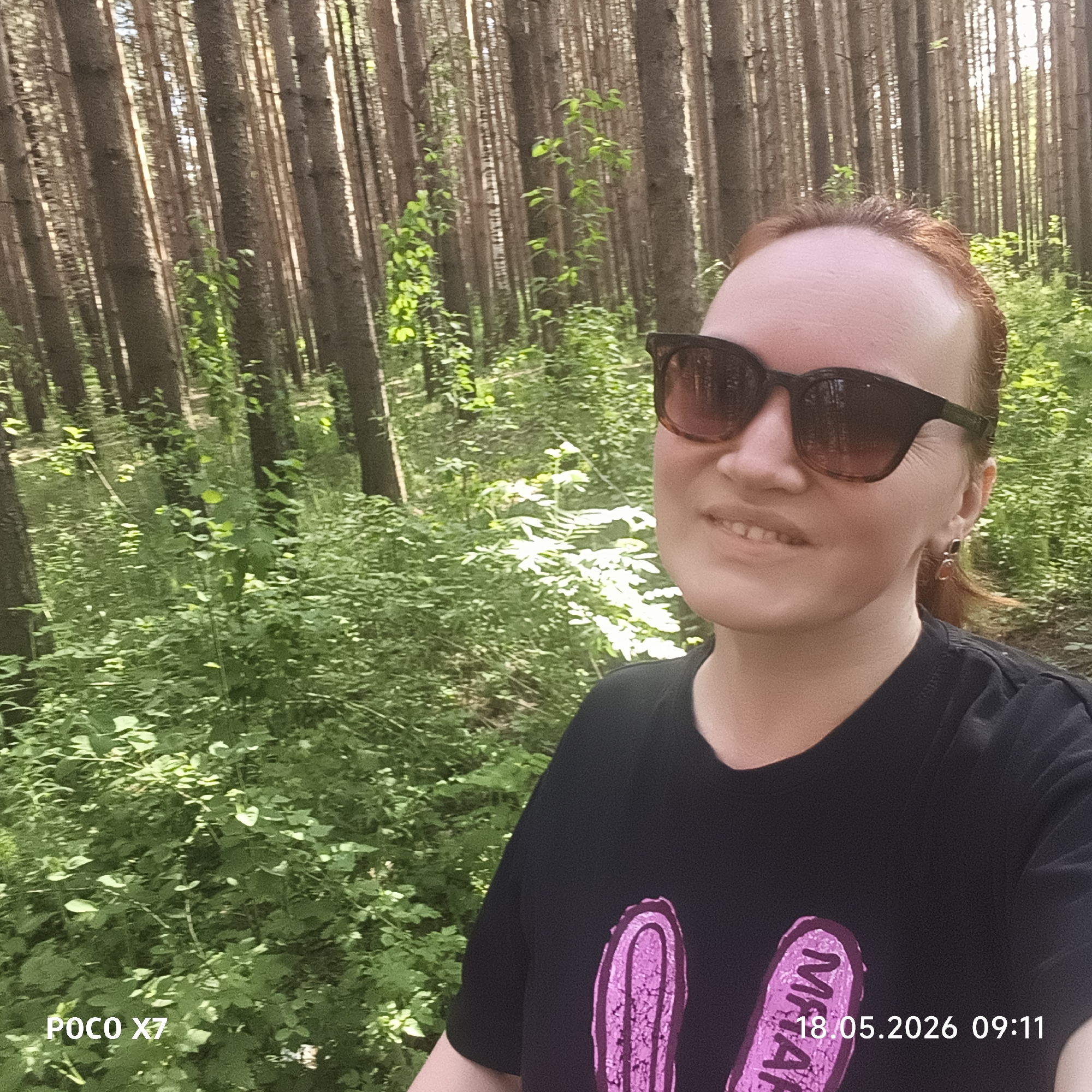 A selfie shows a woman standing in a forest with tall, thin trees. She has reddish hair and is wearing sunglasses and a black t-shirt with a pink design. The ground is covered in green foliage. The photo's timestamp in the lower right corner reads "18.05.2026 09:11". The camera information "POCO X7" is printed in the lower left corner. The location is not provided in the photo.