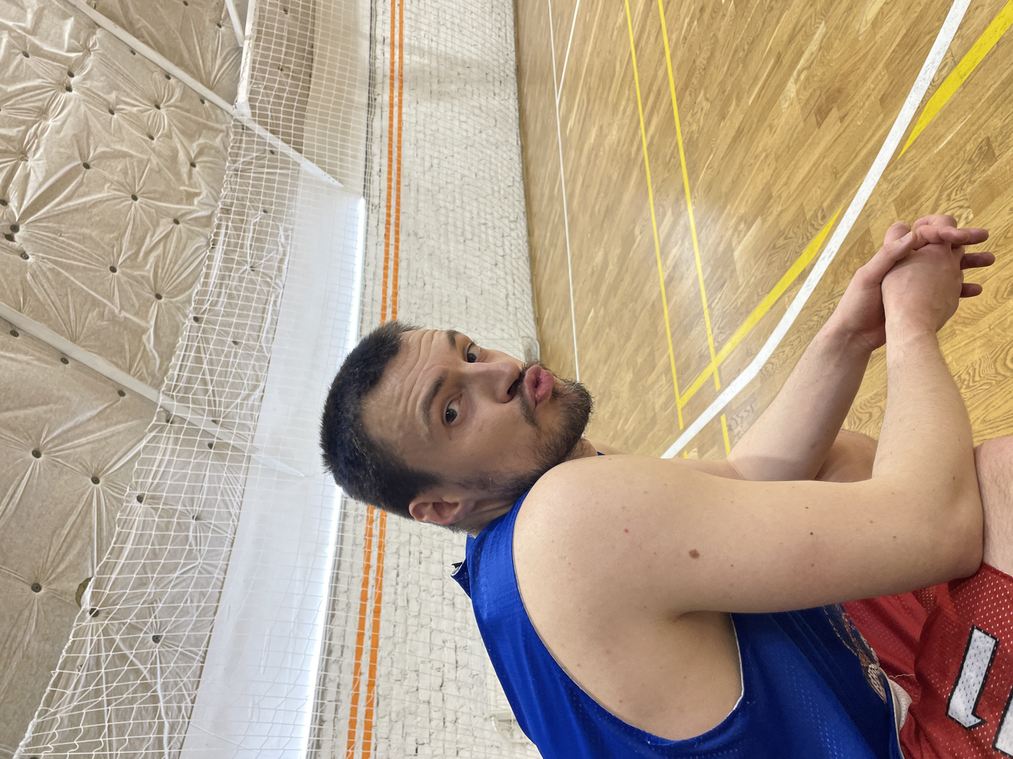 A man with dark hair and a short beard wearing a blue jersey and red shorts is pouting and looking at the camera in a gymnasium. The gymnasium has a wooden floor with yellow and white lines, a white net covering the walls, and a white ceiling with geometric patterns. The photograph was taken inside a gymnasium and there is no way to tell the location in Saint Petersburg, Russia.