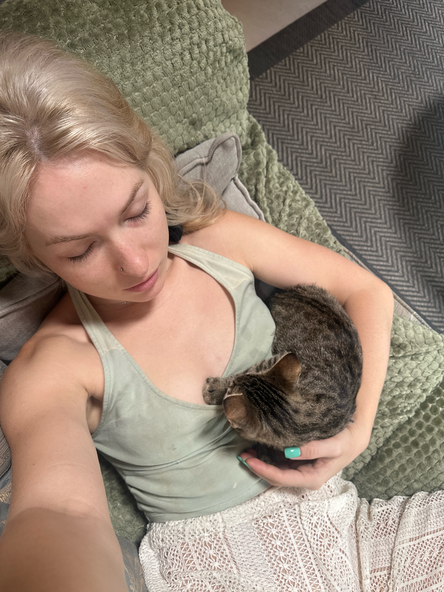 A young woman with blonde hair and closed eyes is lying on a couch, holding a tabby cat in her arms. She is wearing a light green tank top and white lacy shorts. The cat is curled up on her chest, with its head resting on her hand. The couch is covered with a green textured blanket. A herringbone patterned rug is visible on the floor to the right of the couch. The scene appears to be indoors, in a domestic setting, possibly a living room. The woman has a nose ring and her fingernails are painted a bright turquoise color. There is a faint shadow across the lower right portion of the image. The image is taken from a high angle, looking down on the woman and cat. There is no discernible evidence indicating a location in Tbilisi, Georgia.
