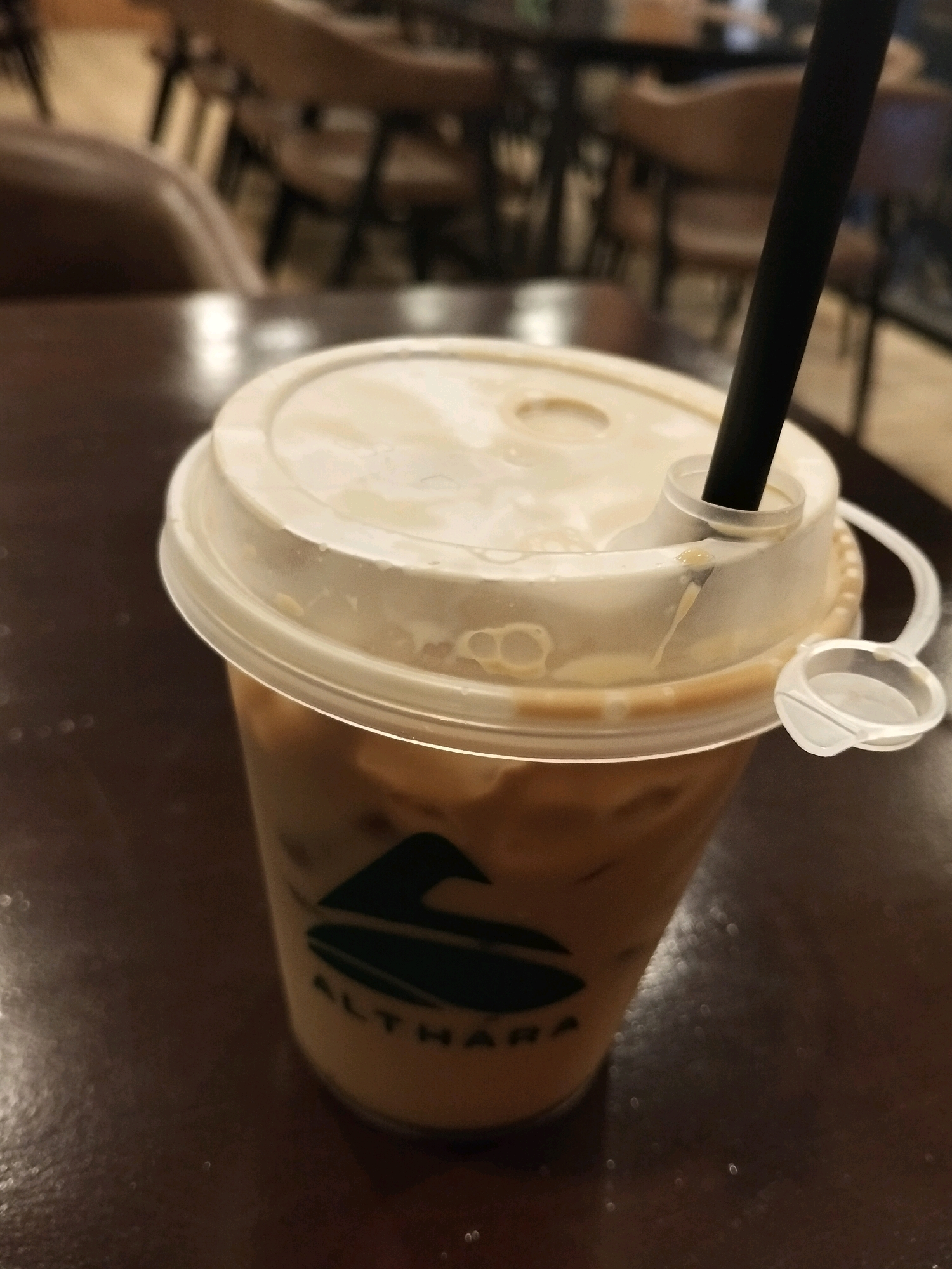 A close-up, slightly elevated, vertical shot focuses on a clear plastic cup filled with a light brown, iced beverage. The cup has a translucent plastic lid with a straw inserted. A white tab is attached to the lid. The cup is positioned on a dark brown wooden table. A dark green logo with stylized leaf shapes and the word "ALTHARA" is visible on the side of the cup. In the blurred background, chairs and tables of a cafe or restaurant are discernible. The lighting is dim, with some reflections on the table surface. The scene is indoors, with no indication of the time of day or specific weather conditions. The location is the city of Arang Limbung, Indonesia.