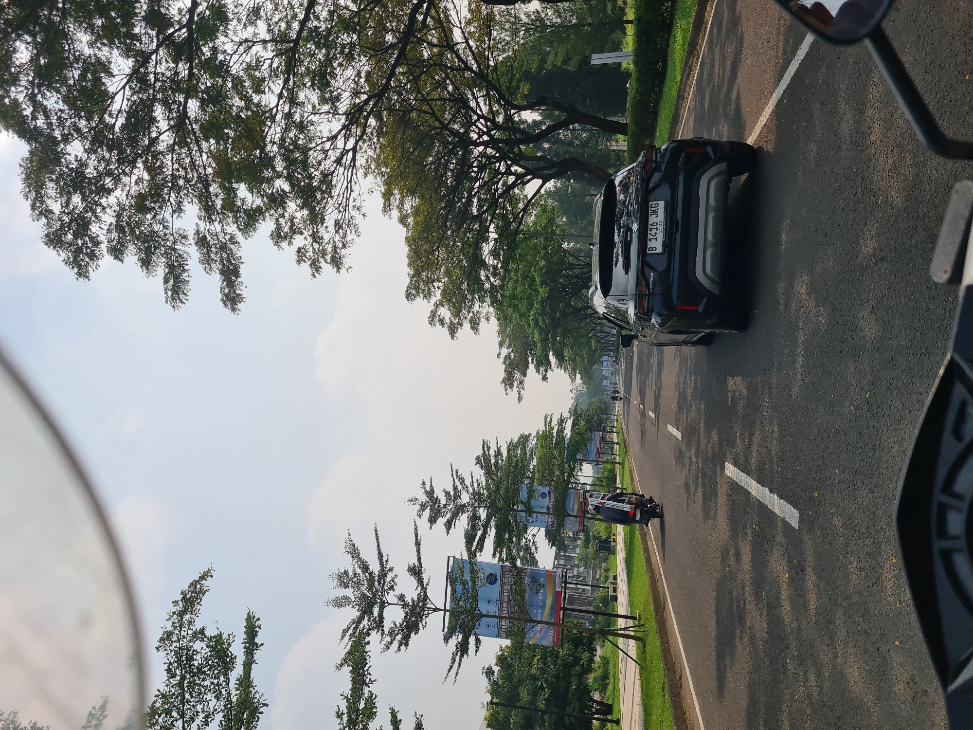 The image captures a daytime scene from the perspective of a moving motorcycle or scooter, showing a multi-lane asphalt road lined with trees and advertising banners. A black car, appearing to be an SUV or crossover, is prominently featured in the right lane, moving away from the viewer. Its license plate, "B 1416 JKG", with an expiry date of "3-20" (likely March 2020 or 2023), identifies its origin as Jakarta, Indonesia.
The road features clear white dashed and solid lane markings. On both sides, mature, leafy trees provide shade, while further down, sparser, possibly younger trees are also visible. Between these trees, several large billboards or banners are erected, displaying graphics and some faint text. While much of the text is illegible due to distance, phrases like "IN THE HEART OF BEAUTY" and a year, possibly "2017" or "2021", can be vaguely discerned on some banners.
The sky is bright and clear with scattered white clouds, indicating fair weather. A green verge and a sidewalk run alongside the road. Another vehicle, possibly a small car or motorcycle, is visible further in the distance on the road. Part of a rearview mirror or handlebar is visible in the upper right foreground, confirming the viewpoint from a two-wheeled vehicle. The scene depicts a busy urban or suburban street environment.
