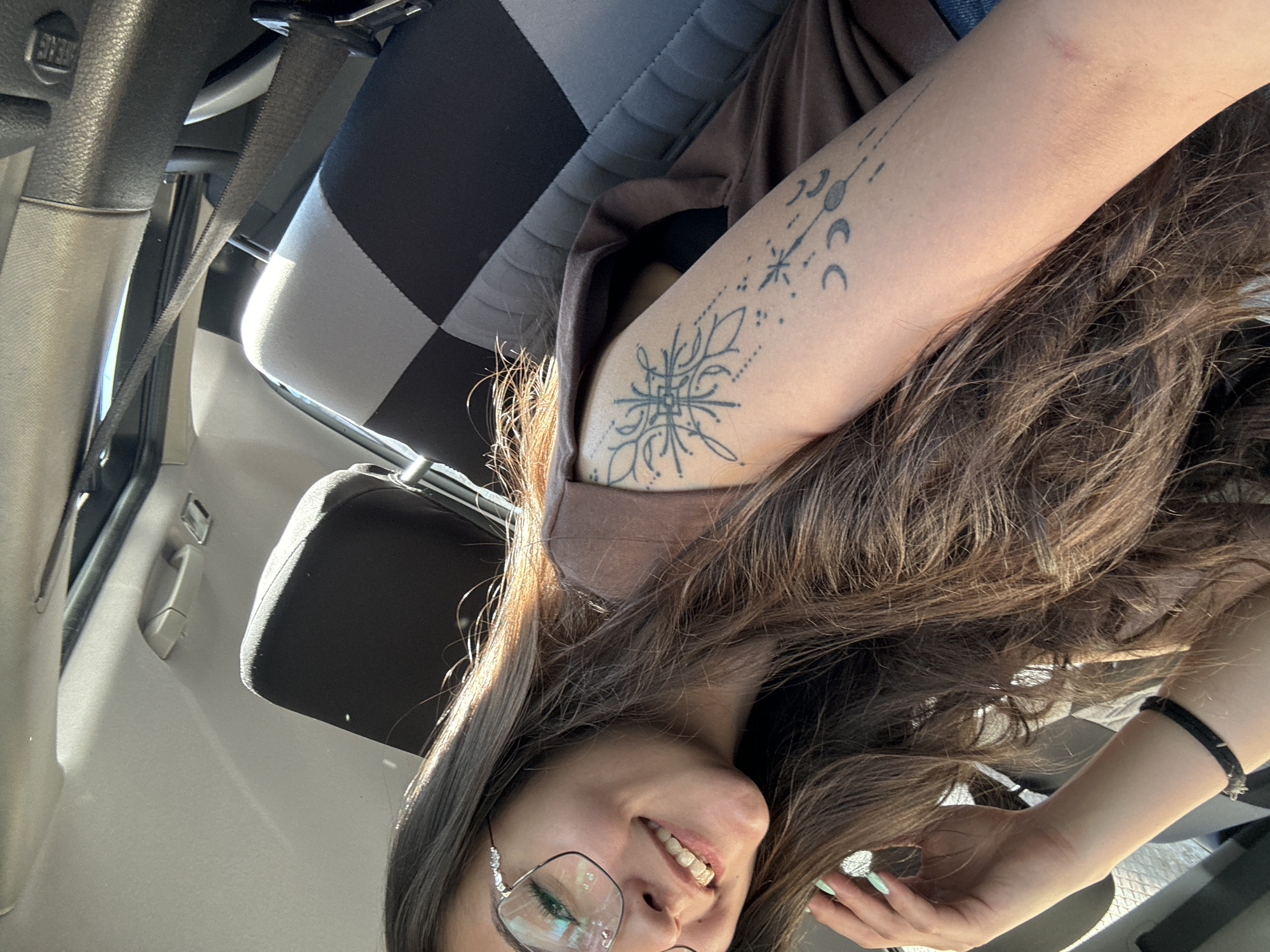 The image depicts a woman in a car. She has long brown hair, wears glasses, and is smiling. She has tattoos on her left upper arm, including a geometric design and lunar phases. The car's interior is visible, including a checkered seat cover, the seat belt, and dashboard elements. The photo was not taken in Dupnitsa, Bulgaria. There is no evidence to suggest it was taken in Dupnitsa, Bulgaria.