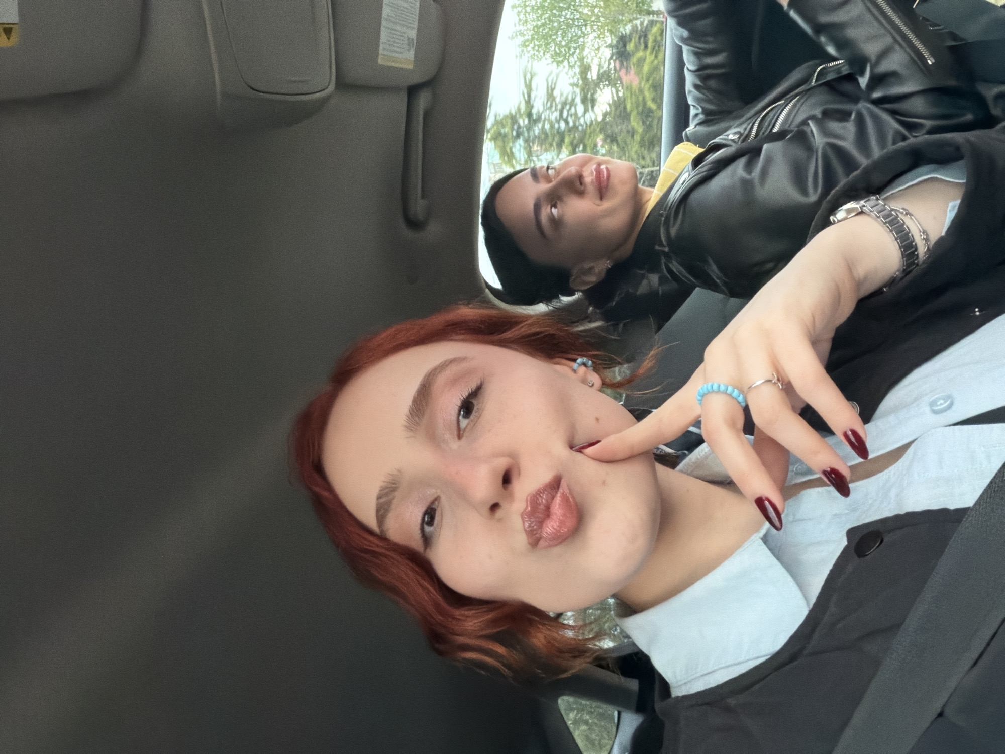 A selfie taken inside a car in Tbilisi, Georgia, features two young individuals during daytime. In the foreground, a person with reddish-brown hair and light skin looks playfully at the camera, pouting their lips and gently touching their cheek with a finger. They wear a light blue collared shirt under a dark jacket, and their fingers display dark red almond-shaped nails, a light blue beaded ring, and a silver-colored ring.
Behind them, another person with dark hair is visible, looking up and slightly to the right with a gentle smile. They are wearing a black leather jacket over what appears to be a yellow top. A wrist watch with a metal band is visible on their left wrist.
The car's interior, including the gray headliner and sun visors with an illegible warning label, forms the immediate background. Through the car window, green trees and foliage are visible, suggesting an outdoor, natural setting and fair weather. The overall impression is a casual, shared moment between the two individuals, conveying a sense of ease and happiness.