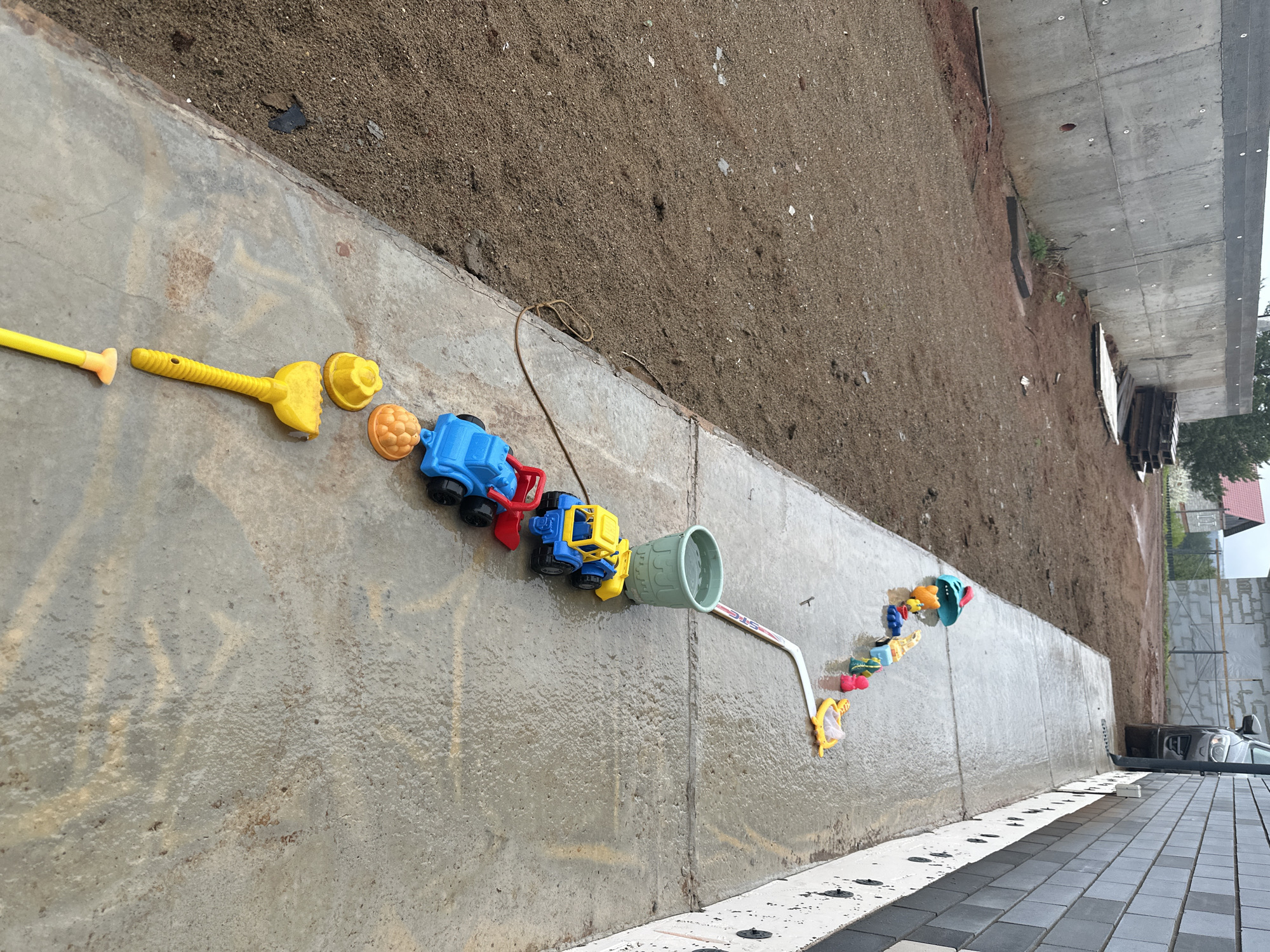 The scene captures a collection of children's beach toys arranged on a concrete surface. From left to right, the objects consist of yellow plastic shovels and molds, a blue and red toy truck, a yellow and blue toy bulldozer, a green pail, and a toy rake and net. A thin rope is seen lying among the toys. The backdrop features a steep earth bank, and, beyond it, there is a structure that appears to be a retaining wall or bridge abutment. In the distance, one can see trees, a building, and a vehicle parked on a paved area. The photograph was taken in Прикамский, Russia.