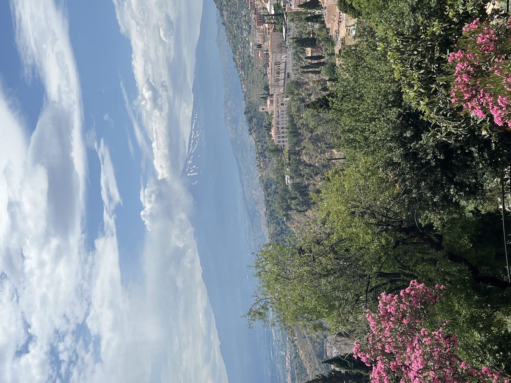 This is a photograph of Mount Etna, a volcano in Sicily, Italy, as seen from Taormina. The volcano is partially obscured by clouds, but its snow-capped summit is visible. In the foreground, there is a lush green landscape with trees and pink flowers. In the middle ground, there is a town with terracotta roofs, perched on a hillside. The sky is a mix of blue and white clouds. The photograph appears to have been taken on a sunny day.