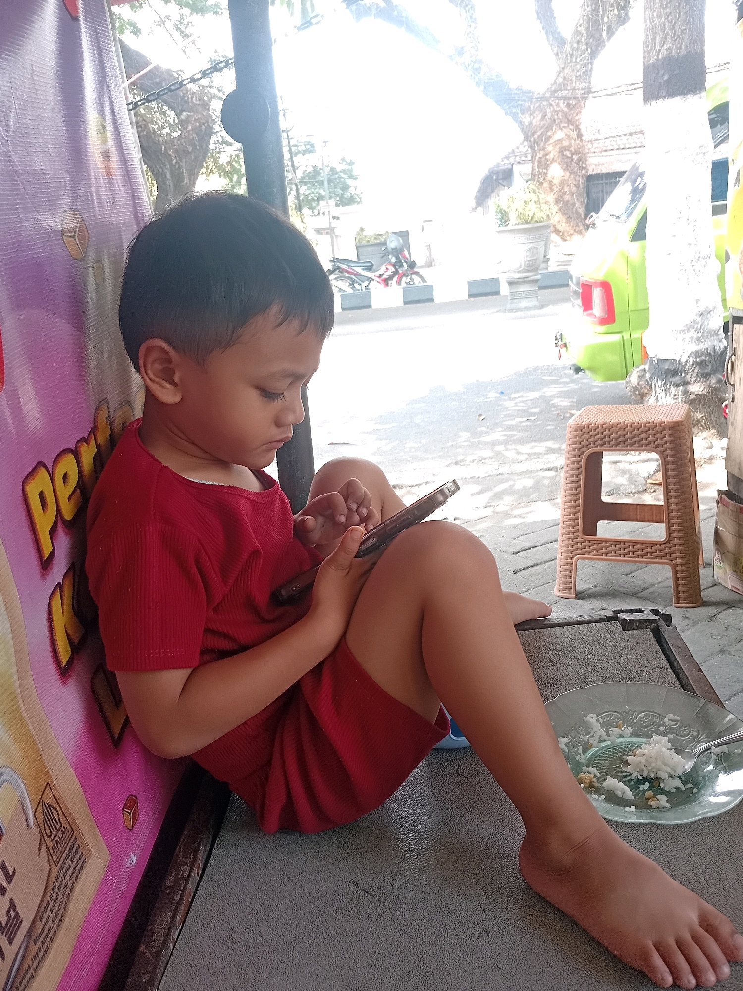 A young boy in a red t-shirt and shorts is sitting outdoors, leaning against a purple banner that says "Pertamina". He is looking down and holding a smartphone in his hands. In front of him, on a grey surface, is a plate of white rice with some green vegetables and a fork. To his right, there is a light brown plastic stool. In the background, there is a large tree, a parked light green vehicle, and a street scene with other parked vehicles and buildings. The image appears to be taken during the daytime, with bright sunlight creating shadows. The setting is likely a public area in Malang, Indonesia, given the Pertamina banner which is a state-owned oil and gas company in Indonesia.
