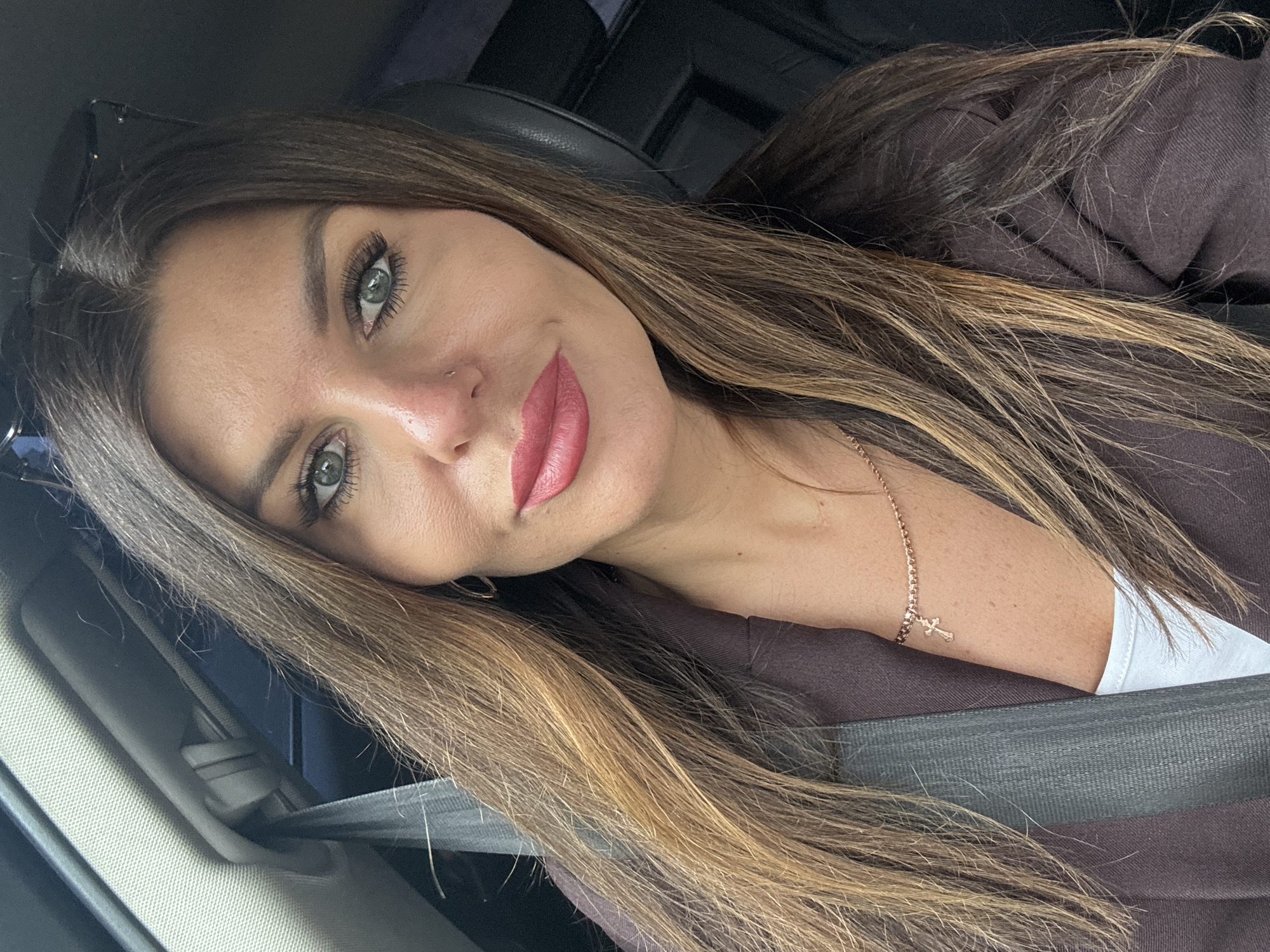 A close-up selfie shows a woman with long, dark brown hair with blonde highlights, wearing a dark brown blazer and a white top. She has green eyes, prominent cheekbones, and full, pink lips. A delicate gold necklace with a cross pendant is visible on her neck, and a thin gold bracelet with a similar cross charm is on her wrist. She is seated in the driver's seat of a car, with a gray seatbelt across her chest. The interior of the car is visible in the background, including a black headrest and a gray headliner. The image appears to have been taken in Chaikovsky, Russia, given the mention of the city.
