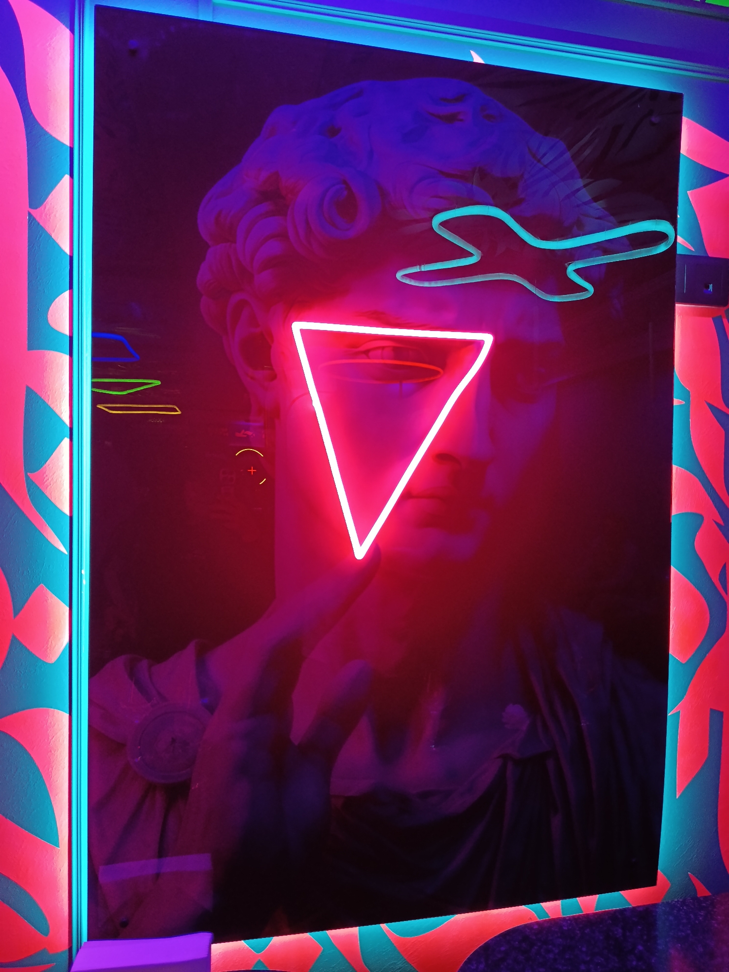 The image shows an indoor scene featuring a framed display with a black background. A print of a classical sculpture, possibly David, is visible behind neon lights in the shape of a triangle (red) and an abstract airplane or shape (turquoise). There are also smaller, horizontal neon lines of yellow, green, and blue. The frame of the display is lined with turquoise neon lighting, and the wall surrounding it is decorated with a repeating pattern of geometric shapes in pink and turquoise. The location is indicated as Unknown, Unknown.
