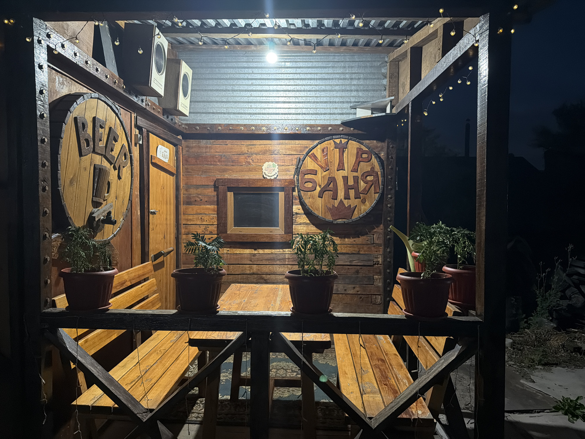 The image depicts an outdoor wooden seating area, possibly a bar or relaxation spot, at night. The structure is made of rough-hewn wood, with visible beams and planks, suggesting a rustic or homemade construction. String lights with small bulbs illuminate the scene.
Dominating the scene are two large circular wooden signs. The one on the left features the word "BEER" in large, carved wooden letters. The sign on the right has Cyrillic lettering that reads "УТР БАНЯ," which translates to "Morning Bath." Below this text is a carved crown.
In front of the wooden wall with the signs, there are wooden benches and tables. Several potted plants are placed on a railing above the benches, adding a touch of greenery. To the left, a door is visible. Two speakers are mounted on the wall above the "BEER" sign. A metal-ribbed roof is visible in the background.
The overall impression is of a cozy, possibly informal, establishment, designed for outdoor leisure. The presence of the "Morning Bath" sign suggests it could be part of a banya (sauna) complex or a recreational area associated with one. The setting appears to be in Semey, Kazakhstan, as indicated by the prompt, though the image itself does not contain any explicit geographical markers beyond what is stated. The night setting and artificial lighting create a warm, intimate atmosphere. There are no people visible in the image.