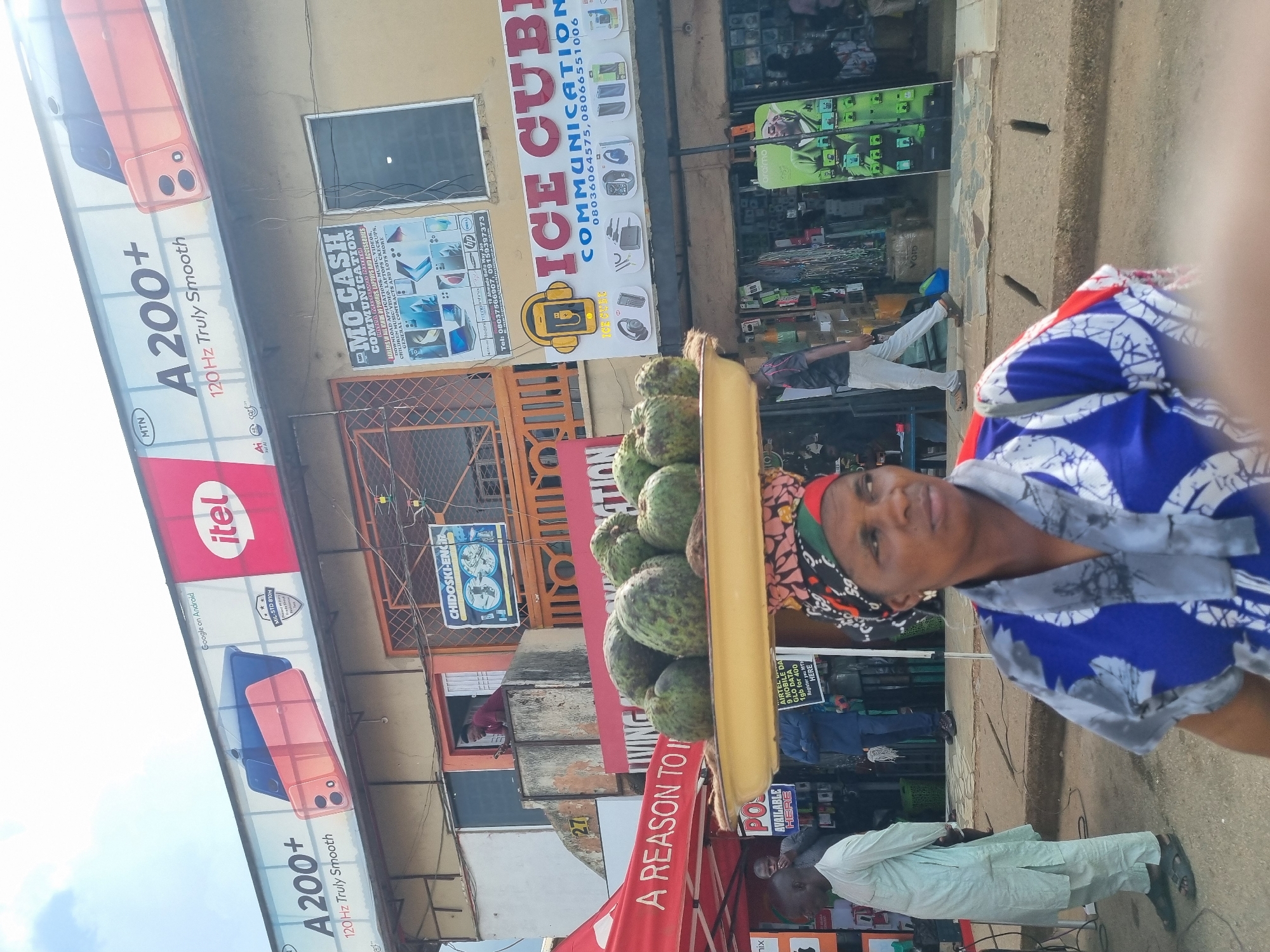 In Jos, Nigeria, a woman with a headwrap and a blue and white patterned top balances a large tray of soursop fruit on her head. She stands in the foreground, looking to her right. Behind her, men in light-colored clothing walk by. Shops line the street, with colorful signage advertising electronics and communication services. A large billboard for "A 200+" phones is visible above the shops. The scene depicts a busy street market environment in the city.