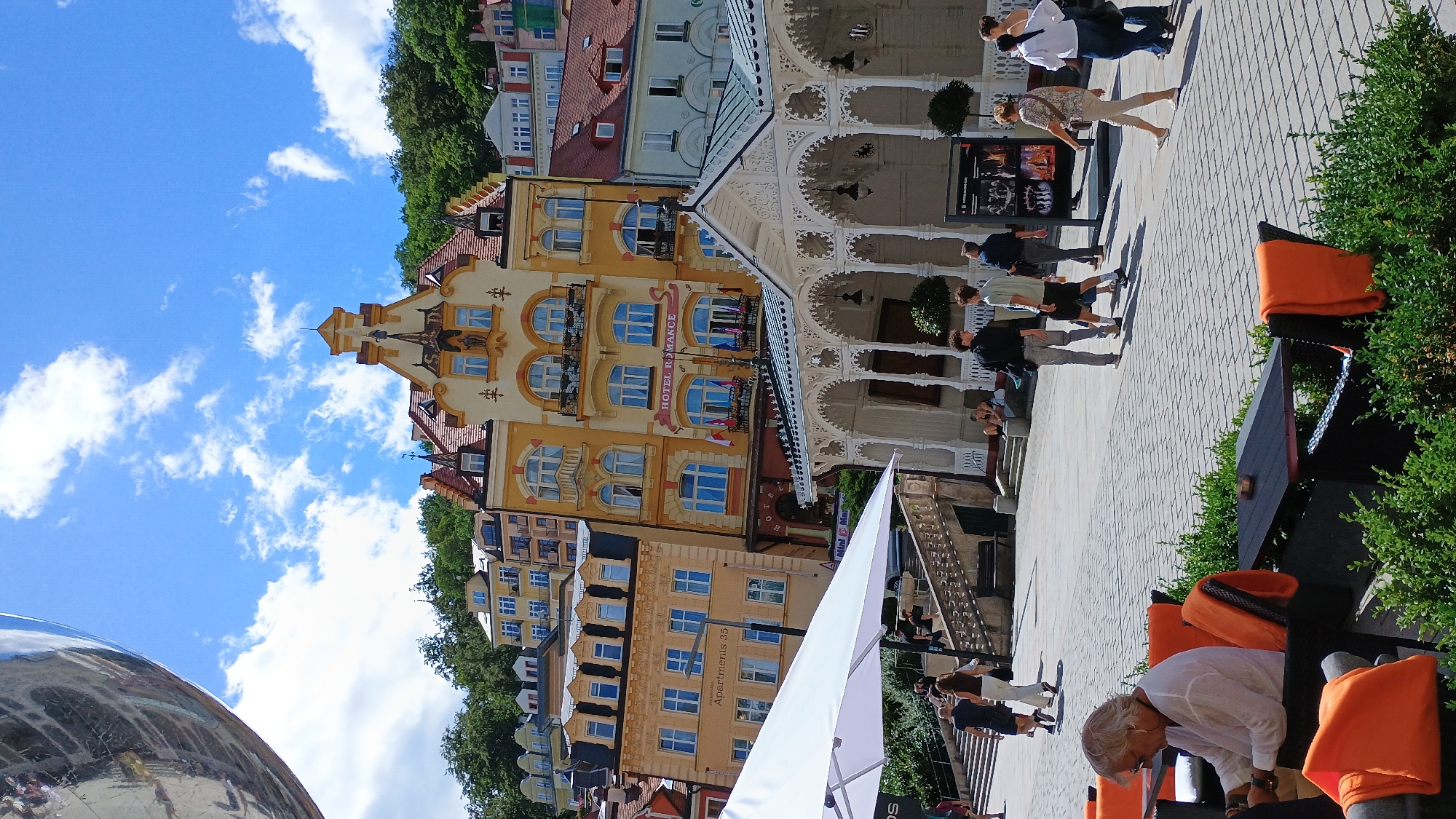 The image depicts a sunny day in Karlovy Vary, Czechia, specifically in the spa town's colonnade area. In the foreground, a woman with grey hair and sunglasses is seated at a table with orange cushions, looking down at something in her hands. Adjacent to her are more tables and chairs, some with orange blankets draped over them, and dense green foliage.
The middle ground features a paved plaza where several people are walking. On the left, a white umbrella shades an area. A white, ornate colonnade with arched openings and decorative metalwork is a prominent feature, with lights hanging from its ceiling. Behind the colonnade, several buildings are visible.
The architecture is colorful and varied. A tall, yellow building with a red roof and elaborate gables stands out. It features multiple balconies and large arched windows. A sign on this building reads "HOTEL ROMANCE". To its left is a lighter yellow building with the text "Apartments 35" visible. Further back, other buildings in shades of yellow, white, and red, with various rooflines and window arrangements, are stacked on a hillside covered in lush green trees. The sky is bright blue with scattered white clouds.