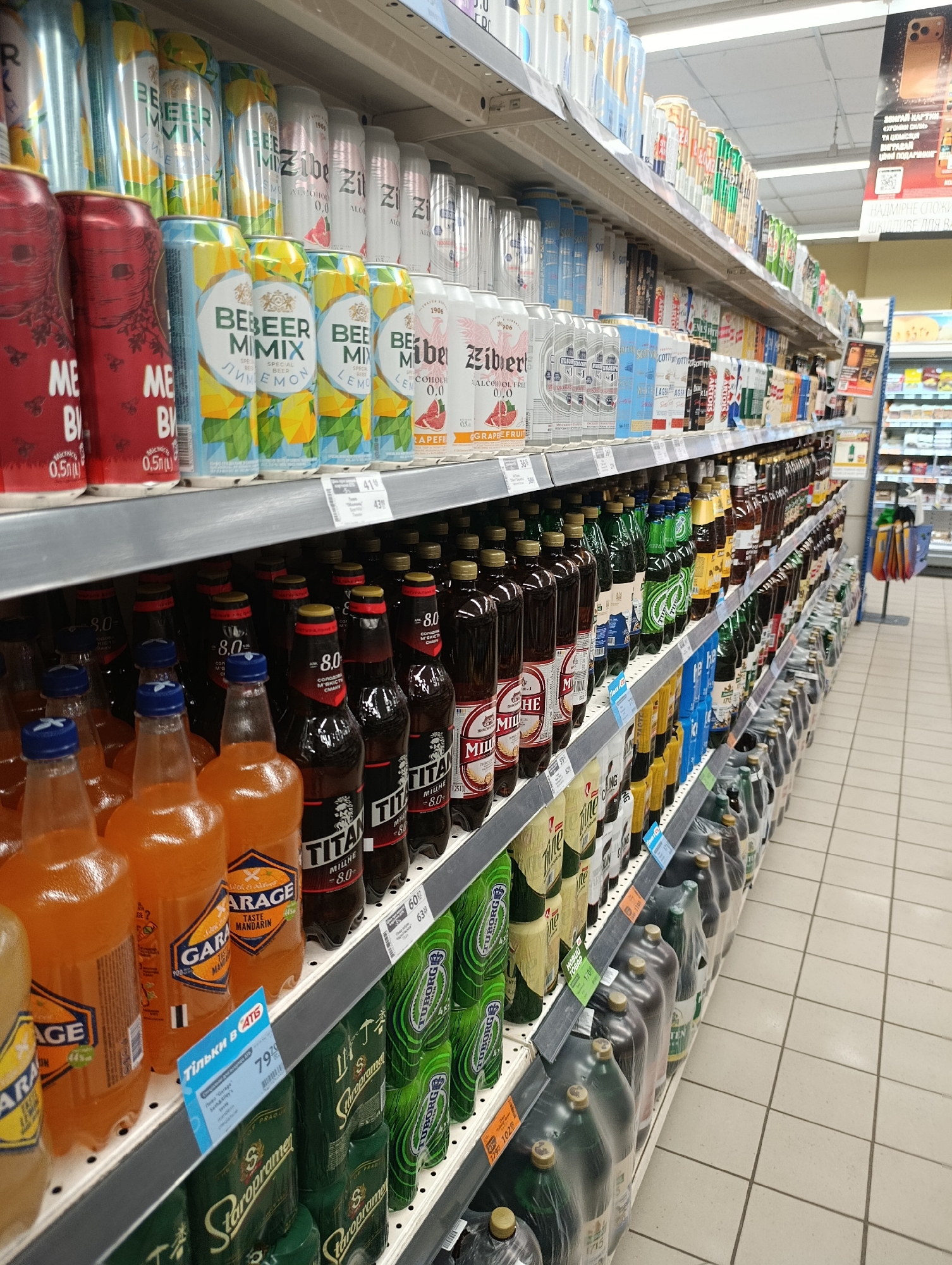 The image shows shelves of beverages in a supermarket in Khorol, Ukraine. The shelves are filled with various canned and bottled drinks, including beer, soda, and other beverages. The drinks are arranged in rows and are mostly colorful, with labels and branding visible. Some of the labels are in Ukrainian. The supermarket appears to be well-stocked and organized. The lighting is bright, typical of a retail environment, with fluorescent lights overhead. The floor is tiled with a light-colored ceramic tile. In the background, other shelves and aisles of the supermarket are visible, along with some shopping carts. There are no people prominently featured in the image, suggesting a focus on the products for sale.