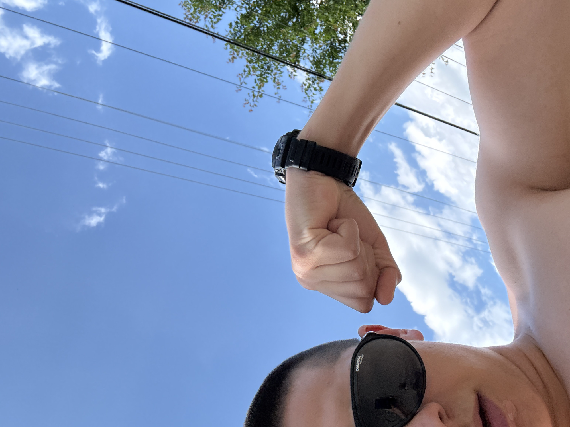 The image is a low-angle outdoor shot, primarily featuring a partially visible person against a blue sky. The person, who has short dark hair, is seen from the nose down, wearing dark Carrera polarized sunglasses. Their bare left shoulder and chest are visible, and their left arm is strongly flexed, showcasing a tensed bicep. A black, robust digital watch is worn on their left wrist. The background consists of a clear blue sky with scattered white cumulus clouds. Multiple parallel utility lines diagonally traverse the upper half of the frame from left to right. On the far right, a small portion of green foliage with reddish-brown branches is visible. There are no identifiable landmarks or signs to determine a specific city or country.