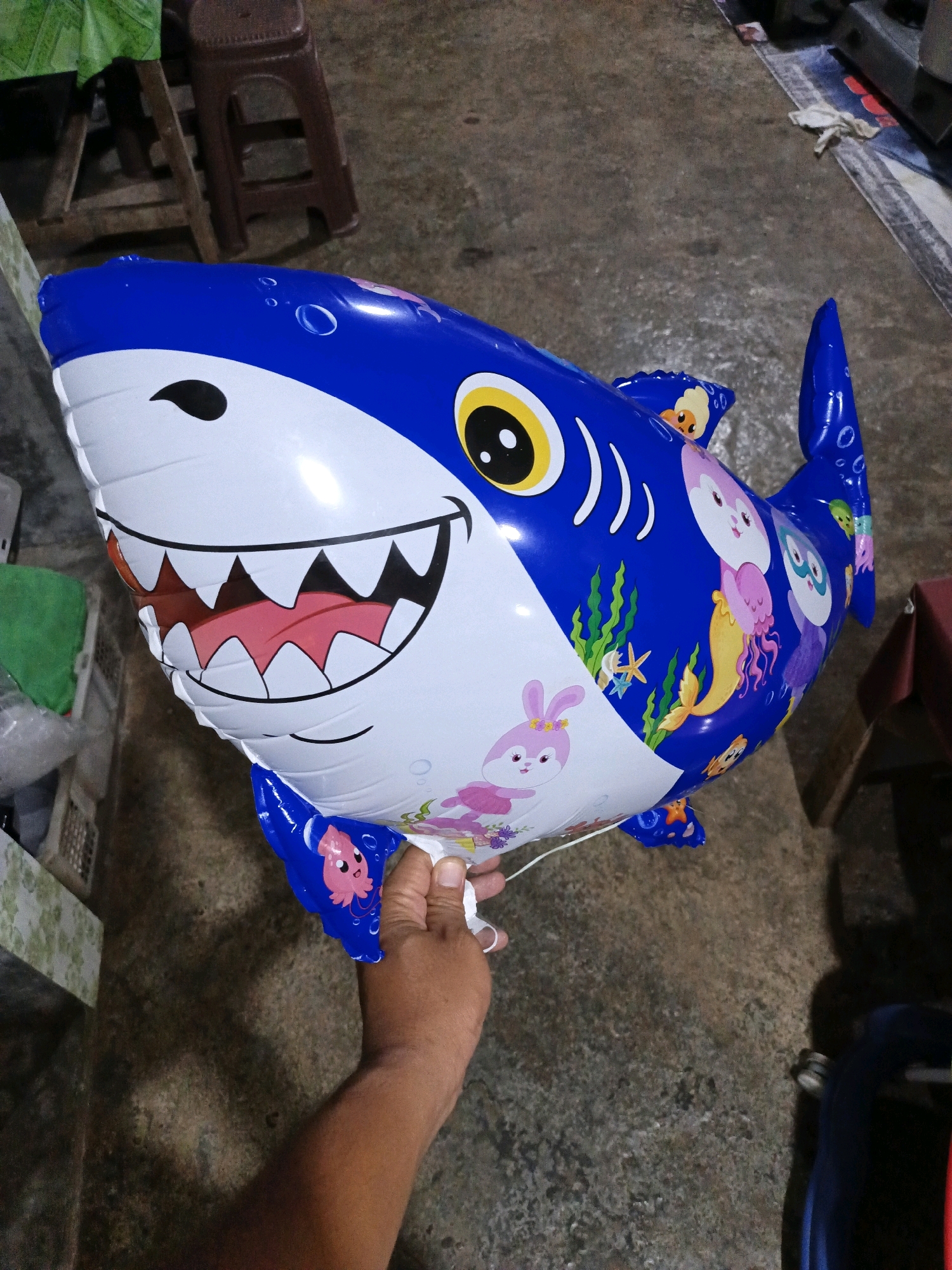 A blue and white inflatable shark balloon is held by a person's hand in Keleyan, Indonesia. The balloon is decorated with cartoon sea creatures, including a pink rabbit, a jellyfish, and a squid. The background consists of a concrete floor, a wooden stool, and a green plastic covering. The lighting is natural and the scene is captured from a slightly elevated angle.