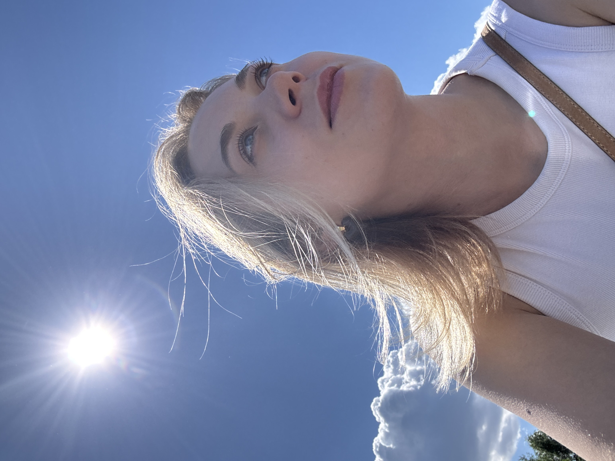 A low-angle, upward-facing photograph captures a person with fair skin and blonde hair against a blue sky with the sun visible. The person is wearing a white tank top with a brown strap across their shoulder. The sun is positioned to the left of the frame, and white cumulus clouds are partially visible. The scene is most likely not within the city of Skulte in Latvia because this photograph does not contain definitive features that would link it to that location.