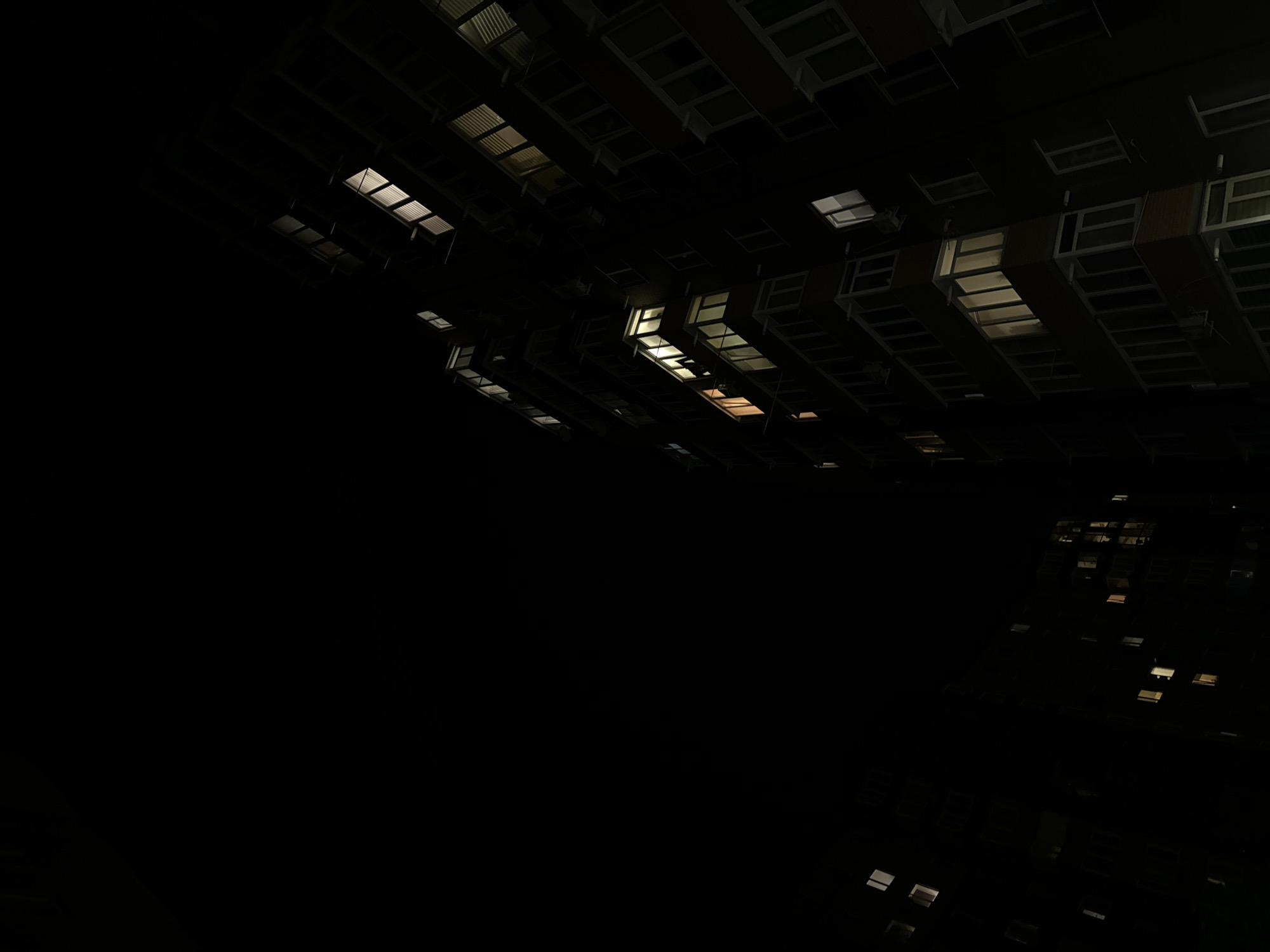 The media file depicts an abstract, dimly lit view of what appears to be the exterior of a large building or complex at night. The perspective is angled upwards, revealing rows of illuminated windows and architectural elements against a very dark background, suggesting a significant portion of the scene is unlit. The illuminated windows suggest interior lighting, with some emitting a warm, yellowish glow and others a cooler, white light.
The overall impression is one of a vast, dark structure with scattered points of light. There are no visible people or overt activities. The scene's low light conditions and the abstract framing create a somewhat mysterious or even desolate atmosphere. The orientation of the windows and the lines of the building suggest a somewhat angular or layered architectural design.
The provided location context of Vinnytsia, Ukraine, does not offer specific visual cues within the image itself. The time of day is clearly night due to the darkness and the artificial illumination from the windows. There are no discernible weather conditions. No text is visible in the image. The focus is on the interplay of light and shadow on a large, man-made structure.
