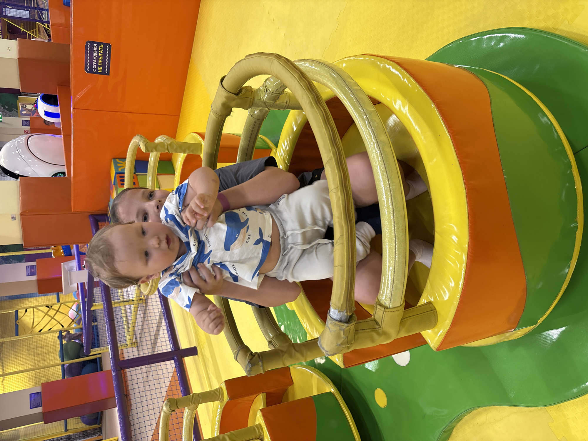 Two children are playing in an indoor playground. One child is holding a baby, who is wearing a white shirt with blue whales and white pants. The older child is wearing a grey shirt. They are inside of a playground tunnel with yellow, orange, and green padded sides. The surrounding play area has orange, yellow, and purple elements. A sign is visible, written in Russian. The location is identified as the city of Izhevsk, Russia.