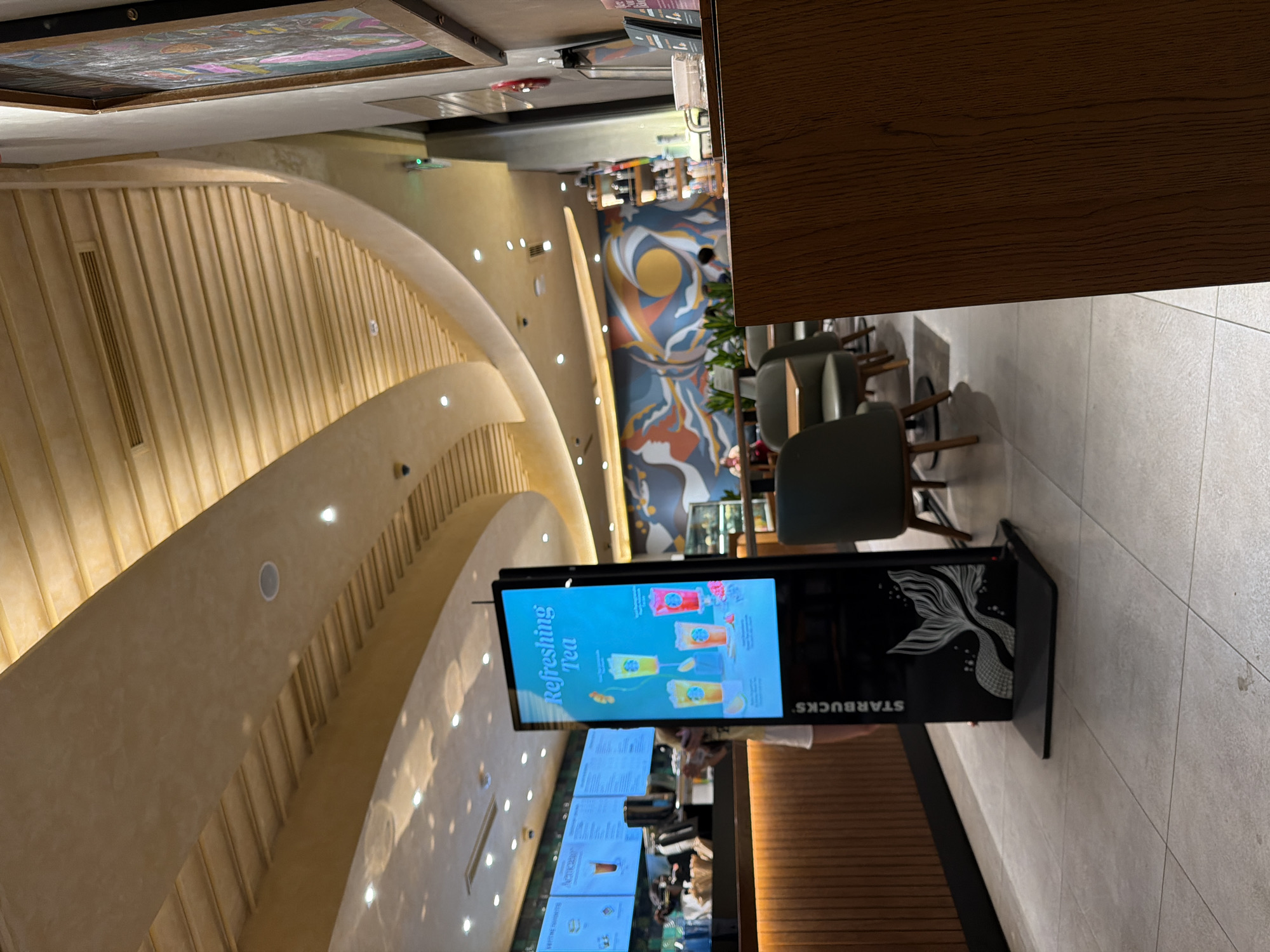 The image captures the interior of a Starbucks store in Da Nang, Vietnam, as indicated by the text on the digital menu board. The scene is illuminated by warm, ambient lighting. A prominent digital display showcases refreshing teas, with graphics of beverages and text reading "Refreshing Tea." To the left, a counter area with menu boards and a barista station is visible. The environment features a modern aesthetic with curved ceilings, wooden accents, and tiled flooring. Seating arrangements with tables and chairs are present in the mid-ground, and a colorful mural adorns the back wall. A person is partially visible near the counter. The overall impression is that of a well-lit, contemporary coffee shop setting.