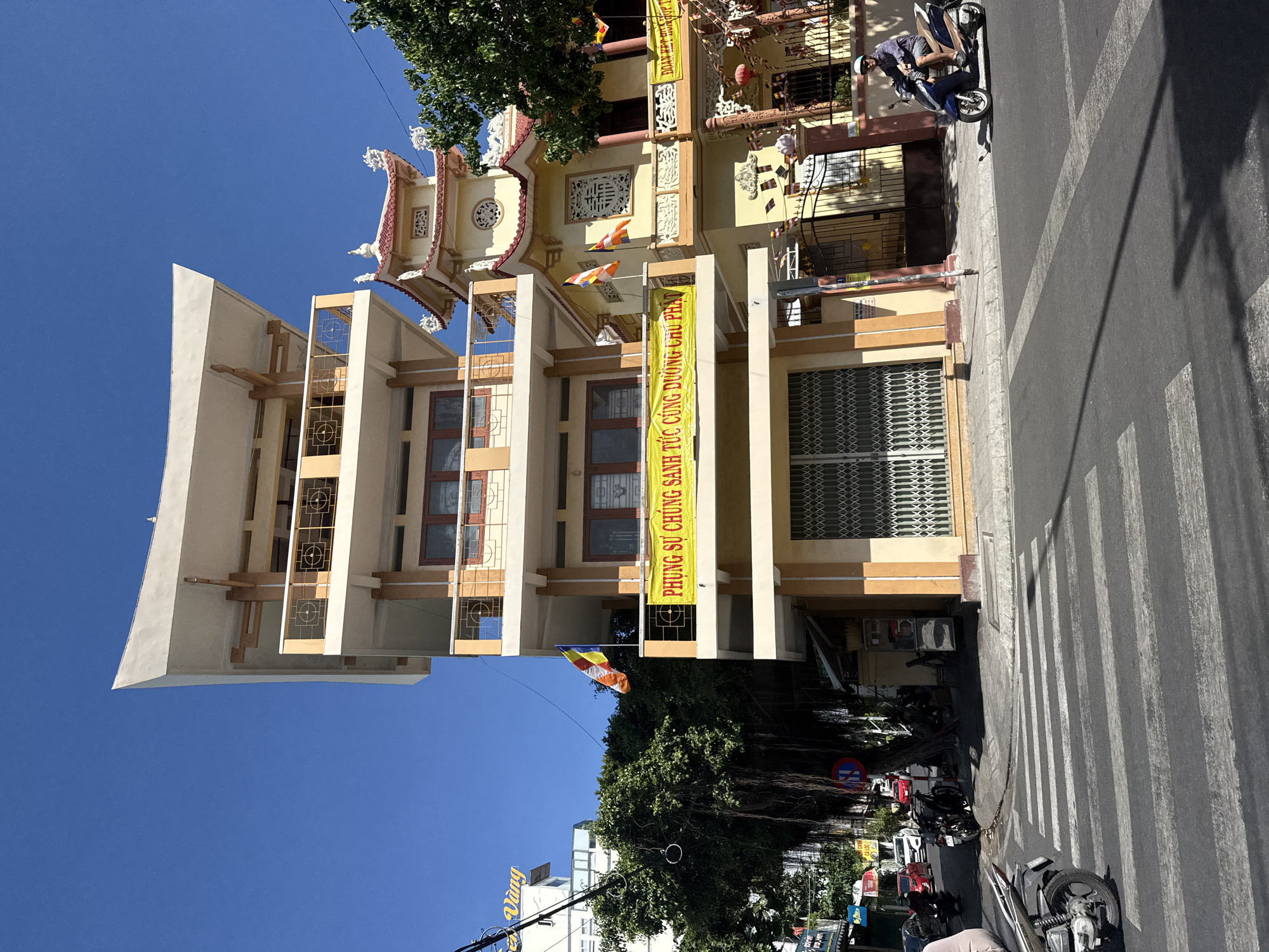 The image shows a multi-story building with Vietnamese architectural design, located in Nha Trang ward, Vietnam. The building has a creamy-yellow facade with decorative elements, including banners with Vietnamese script. The building is flanked by trees. Motorbikes are parked on the street. There are pedestrian crosswalk lines visible on the street. A person is sitting on a motorbike in the foreground. The sky is blue.