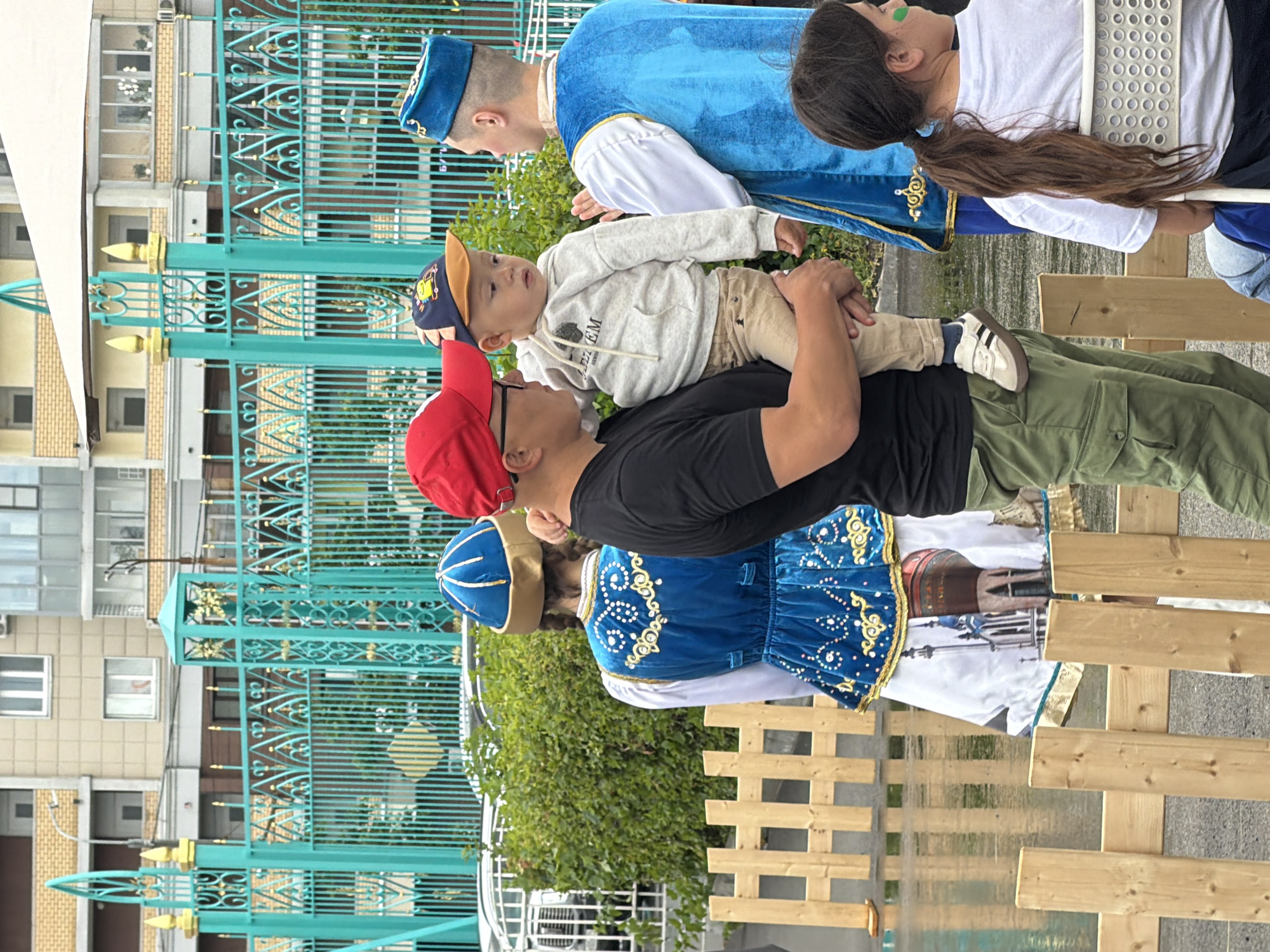 In the city of Unknown, Unknown, a man in a red cap and black shirt holds a young child in a grey hoodie and beige pants. The child wears a blue and orange cap with a cartoon image. To their right, a person in traditional blue and gold attire with a blue cap speaks with the man. Further to the right, another individual in a similar blue and gold outfit stands with their back to the camera. In the foreground, a young girl with a brown ponytail and green face paint sits in a white chair, facing away from the camera. A wooden fence runs along the bottom of the image, and behind the people is a turquoise metal fence with ornate designs, with buildings and greenery visible behind it. The ground appears wet and reflective.