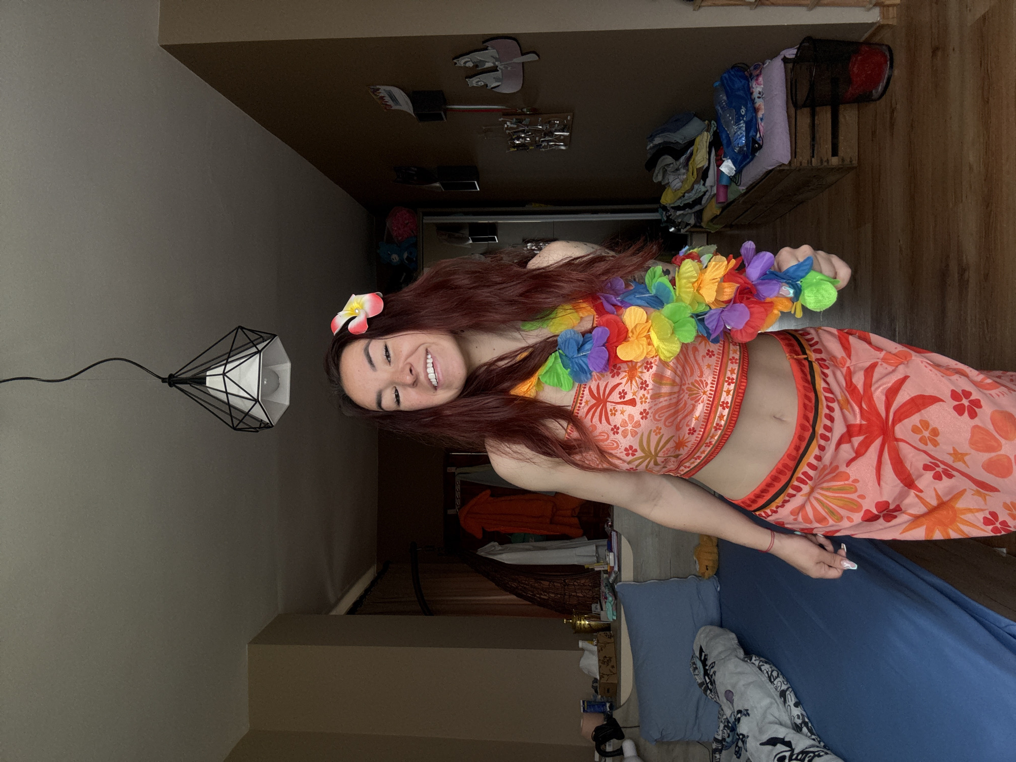 A young woman with long, auburn hair, wearing a Hawaiian-themed outfit consisting of a crop top and skirt with floral patterns, and a colorful lei, stands smiling in what appears to be a bedroom. A flower is clipped in her hair. The room features a bed with blue sheets, a lamp with a geometric shade, and a wardrobe visible in the background. The location is indeterminate.