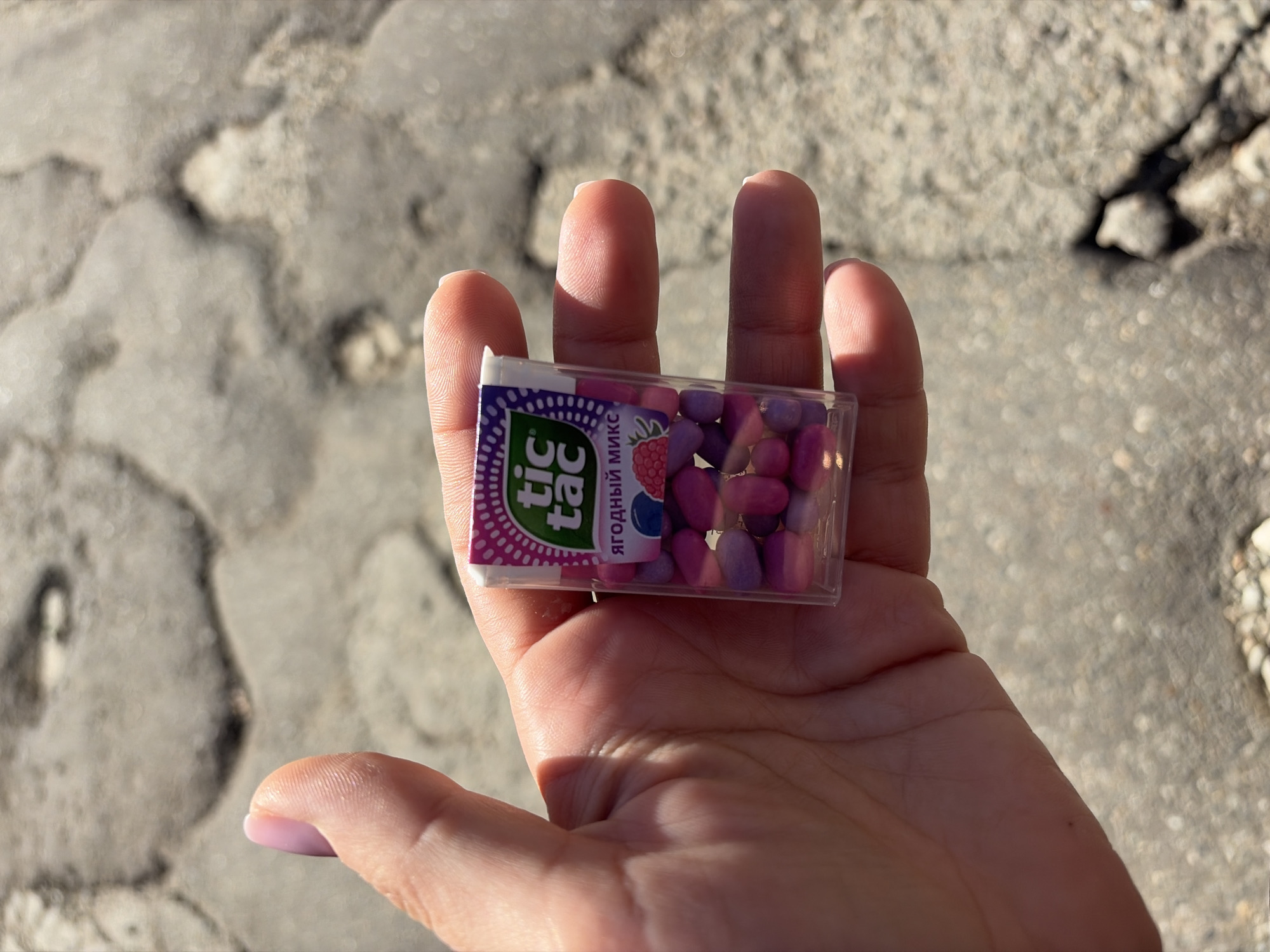 A human hand holds a translucent Tic Tac candy box in a close-up, outdoor shot. The hand, with light pink polished fingernails, is positioned against a textured, light-colored ground surface, likely cracked concrete or asphalt, suggesting an outdoor setting in Sochi, Russia. Bright sunlight illuminates the scene, casting clear shadows from the hand and the candy box onto the palm and the ground.
The Tic Tac box contains numerous small, oval-shaped candies in various shades of pink and purple. The label on the box is predominantly purple and pink, featuring the distinctive green "tic tac" logo. Below the logo, the Russian text "ЯГОДНЫЙ МИКС" (YAGODNYY MIKS), meaning "Berry Mix," is visible, accompanied by small illustrations of a raspberry and a blueberry. The activity depicted is simply the holding of the candy box. The time of day appears to be bright daytime due to the strong shadows.