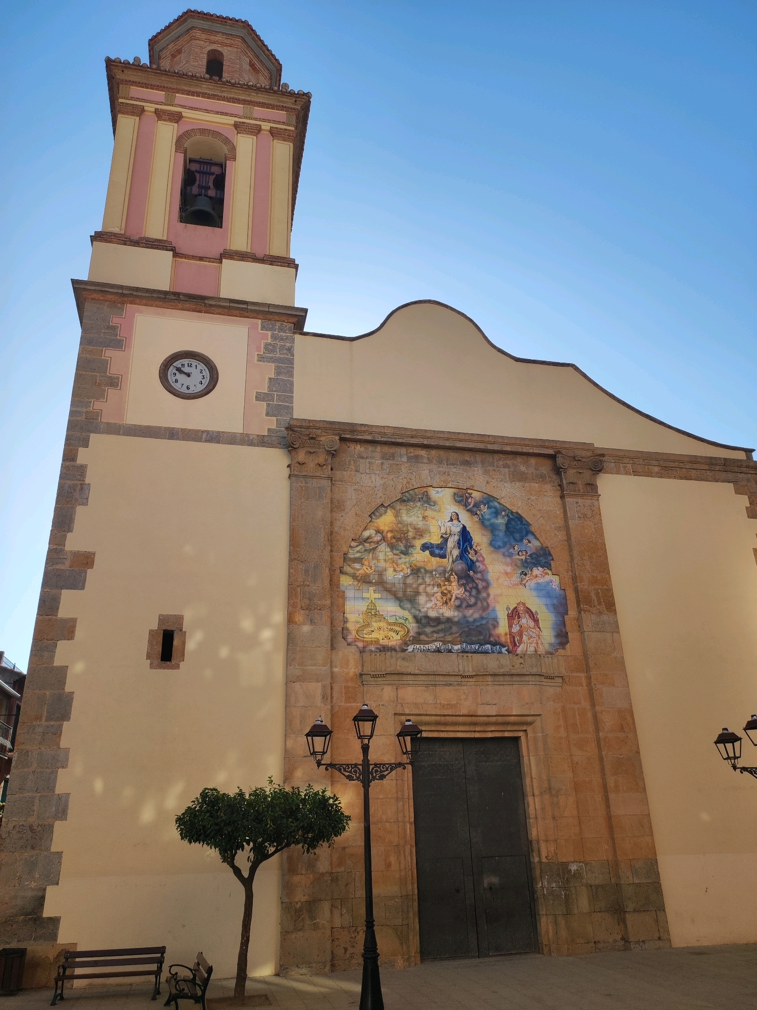 This image depicts the facade of a church in the town of Suera, Spain. The church has a prominent bell tower with pink and yellow accents, and a clock face showing approximately 10:08. Below the clock, there is a small, rectangular window. The main entrance of the church is framed by a large, semicircular tiled mural depicting a religious scene with figures and clouds. Flanking the entrance are decorative stone columns.
In front of the church, a small, leafy tree stands in a paved area. To the left of the tree are two wooden benches, and a trash receptacle is visible behind the left bench. Two black lampposts with multiple lamps are positioned on the right side of the image. The sky is clear and blue. The architecture suggests a Mediterranean or Spanish style.