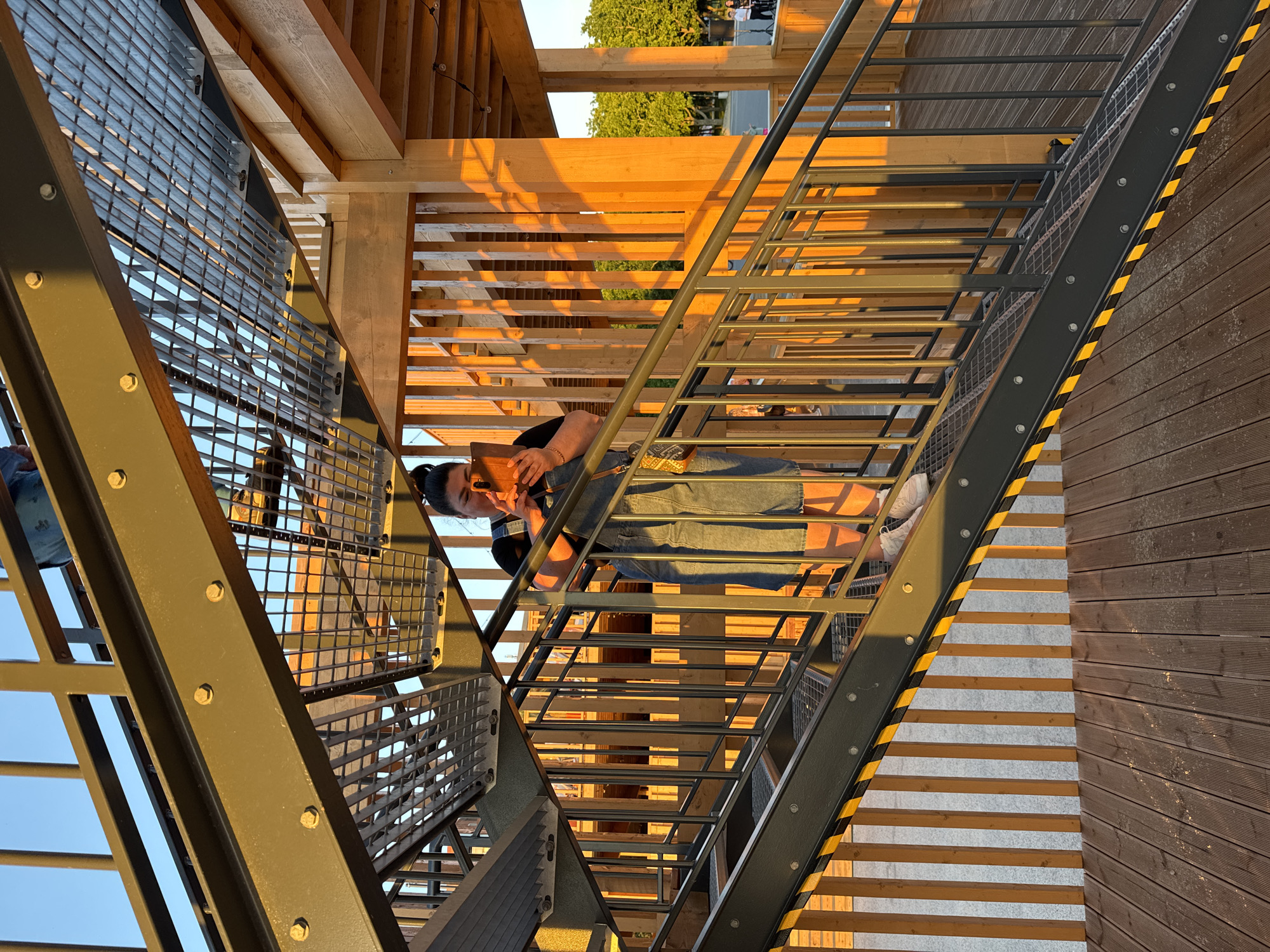 In Tallinn, Estonia, a person is standing on an outdoor staircase of a modern wooden structure, possibly a public art installation or viewing platform, with a sunset glow casting warm light. The person, viewed from the waist down, is wearing denim overalls and white sneakers, holding a smartphone in their hands and looking down at it. The staircase features dark metal railings with yellow safety stripes along the edges, and metal grate treads. The background reveals a wooden structure with vertical slats and dappled sunlight, suggesting an outdoor public space. Trees and a clear sky are visible beyond the structure, and further in the distance, faint figures of people suggest a lively atmosphere.