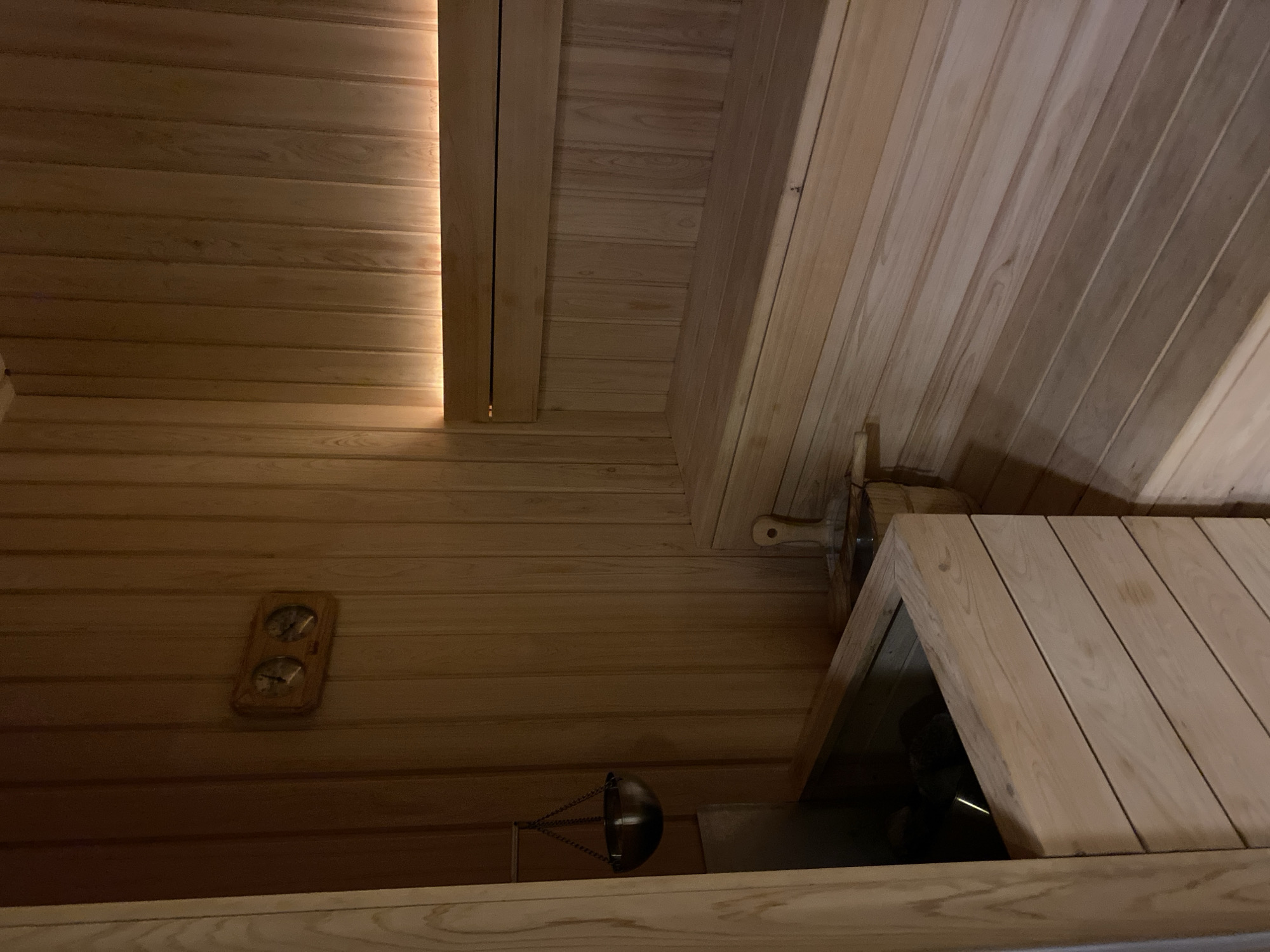 The image depicts the interior of a modern sauna constructed from light-colored wood. The walls and benches are made of vertical wooden planks, with a horizontal bench running along the right side of the image, illuminated by warm lighting at the top. In the background, a thermometer and hygrometer are mounted on the wall. To the left, a metal bucket with a chain hangs over a wooden and metal structure that likely houses the sauna heater. A wooden bucket with a scoop rests on a wooden bench in the middle ground. The scene appears to be empty of people. This sauna is located in Kuta Selatan, Indonesia.