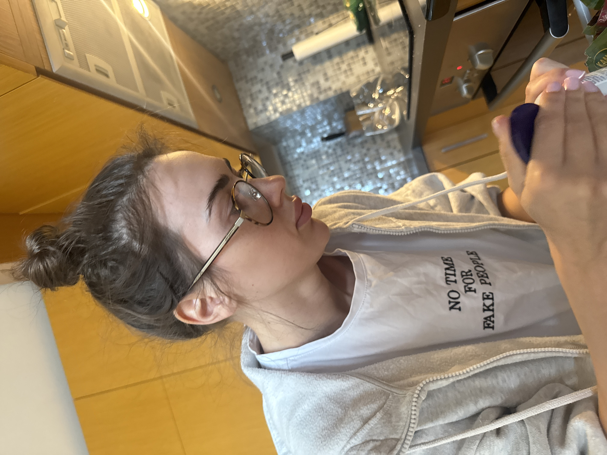 The image captures a close-up, tilted view of a young woman in an indoor setting, likely a kitchen. The woman, with dark hair tied in a bun, is seen in profile, looking upwards and slightly to her right. She wears round, light-colored framed glasses. Her attire consists of a white t-shirt underneath a light grey zip-up hoodie with a drawstring. On the white t-shirt, the text "NO TIME FOR FAKE PEOPLE" is visible.
Her hands, with light pink painted fingernails, are in the foreground on the right, holding a purple object, possibly a spray bottle or container. Her gaze is directed towards this object or something beyond it.
The background reveals typical kitchen elements. Above her, an extractor hood with built-in lighting is visible. Below the hood, a stove or oven with silver control knobs and a glowing red indicator light can be seen. A tiled backsplash with a shiny, possibly mosaic pattern is also present. Wooden cabinets flank the cooking area. Further in the blurry background, a roll of paper (like kitchen roll) on a holder, a green object, and some metallic or glass items are discernible. The lighting appears to be artificial, coming from the kitchen fixtures. The overall scene suggests she is engaged in an activity within the kitchen environment.