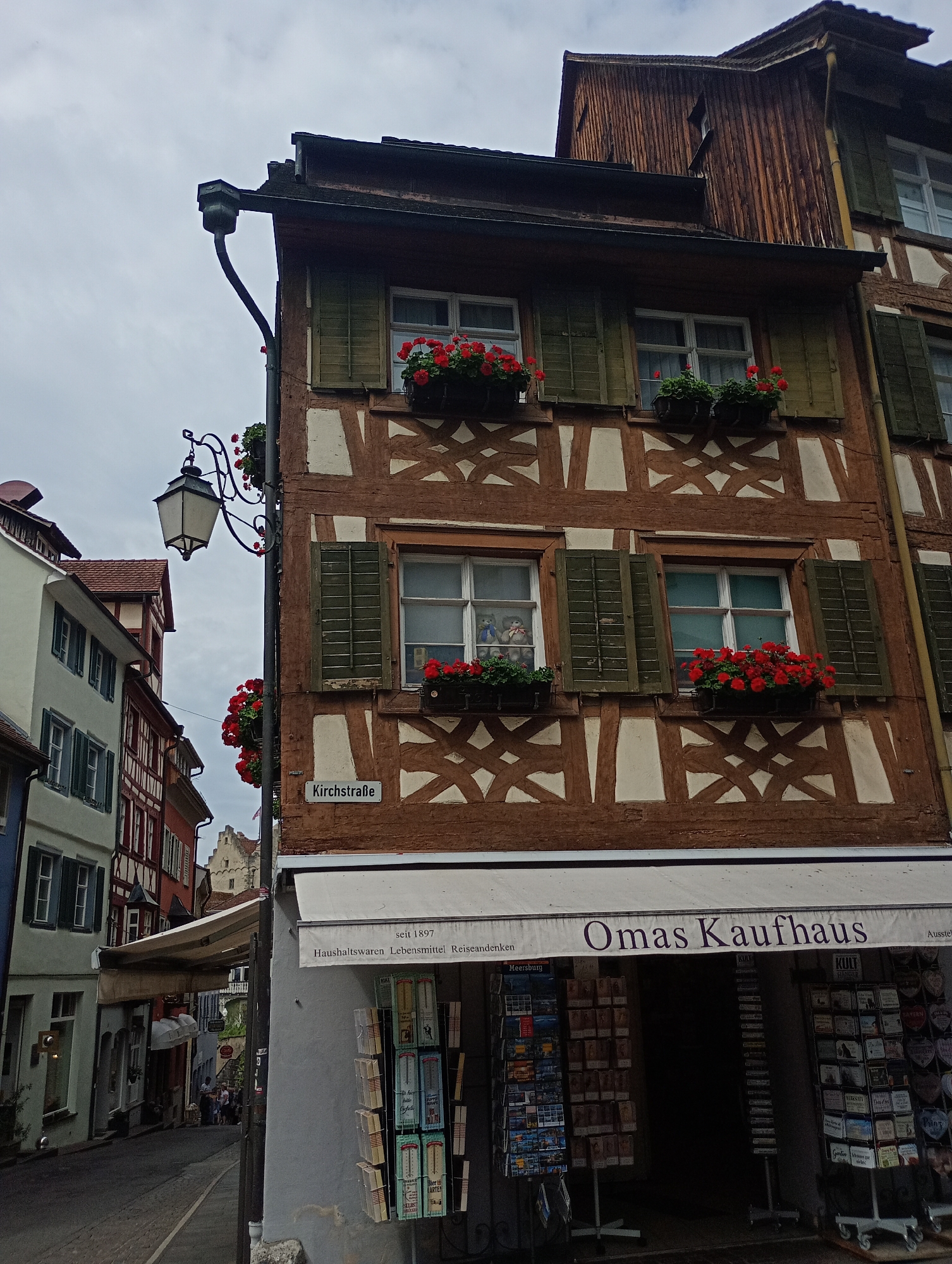 This is a photograph taken in the city of Meersburg, Germany. The image captures a charming street scene with a focus on traditional half-timbered architecture. The central building is a two-story structure with a rich brown timber frame and cream-colored infill. It features several windows with dark green shutters and window boxes overflowing with vibrant red flowers. A street sign reads "Kirchstraße," indicating the street's name.
To the left of the main building, another building with a pale green facade and similar architectural style is visible, along with glimpses of red-tiled roofs and other buildings further down the street, suggesting a historical urban environment. A vintage street lamp with ornate detailing hangs from a pole, adding to the old-world ambiance.
In the foreground, a store with a white awning displays the name "Omas Kaufhaus" in large purple lettering. The storefront showcases various items for sale, including postcards and other small goods displayed on racks outside. The street itself is paved with cobblestones, and the overall atmosphere is one of a picturesque, historic European town. The sky is overcast, contributing to a soft, diffused light.