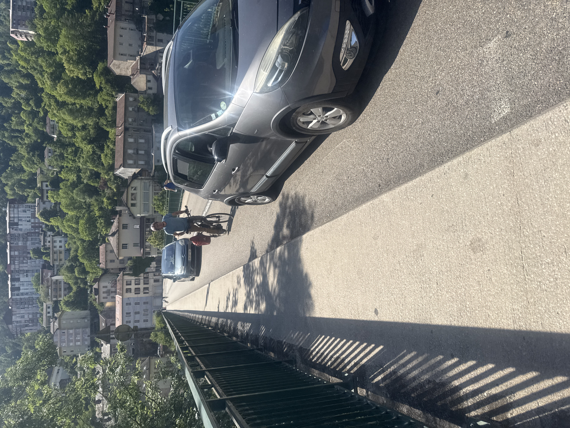 On a sunny day, a cyclist wearing a blue shirt and brown shorts rides a bicycle with a red bag attached to the back. They are on a bridge with a metal railing on the left. A gray car is in front of the cyclist, and another blue car is behind the cyclist. In the background, there are buildings on a hillside covered in green trees. The buildings are a mix of residential and commercial properties, with some appearing to be apartment buildings. The architecture suggests a European setting. The overall scene depicts a street in Saint-Claude, France, with a mix of transportation and urban infrastructure.