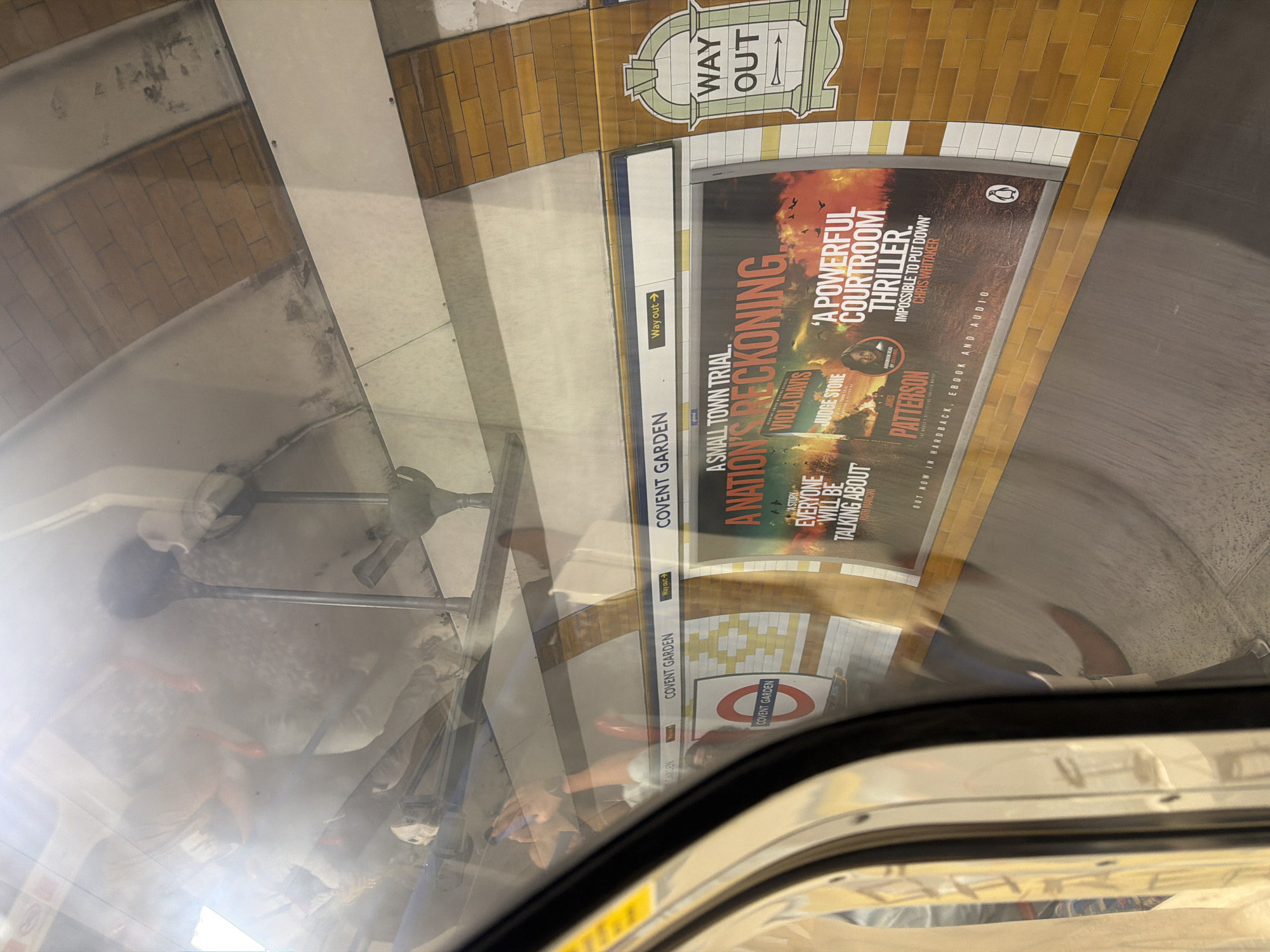 The image shows the interior of a London Underground train carriage. Outside the window, a poster advertising "A Nation's Reckoning" by James Patterson and Viola Davis is visible on the platform. The poster is displayed at Covent Garden station, as indicated by the station name and logo on the platform wall. A "Way Out" sign is also visible. Reflections in the train window show passengers inside the carriage. The walls of the station are tiled with yellow and white tiles.