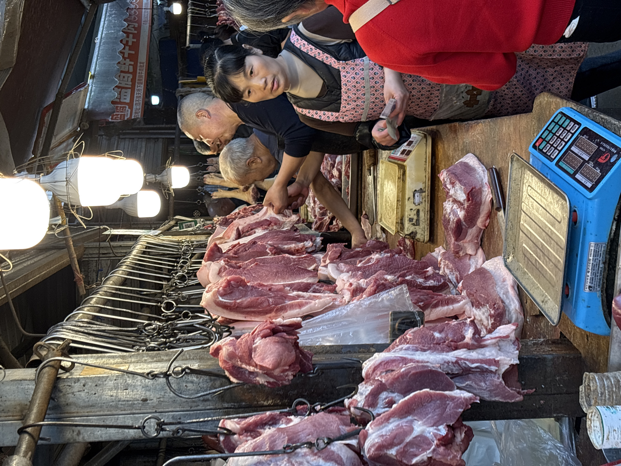 A bustling meat market in Jiangjin, China, features a wooden stall laden with raw cuts of pork. A woman in a pink apron is taking cash from a customer in a red sweater. Two men are seen handling the meat. A blue digital scale sits on the counter, next to another older model. A row of metal hooks hangs above, some holding cuts of meat. Fluorescent lights illuminate the scene. A sign with Chinese characters hangs above the stall.